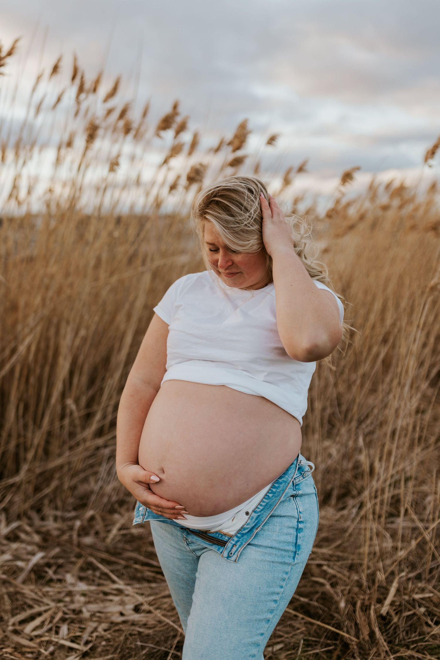 Pregnant woman in white crop top and jeans holding baby bump in golden field.