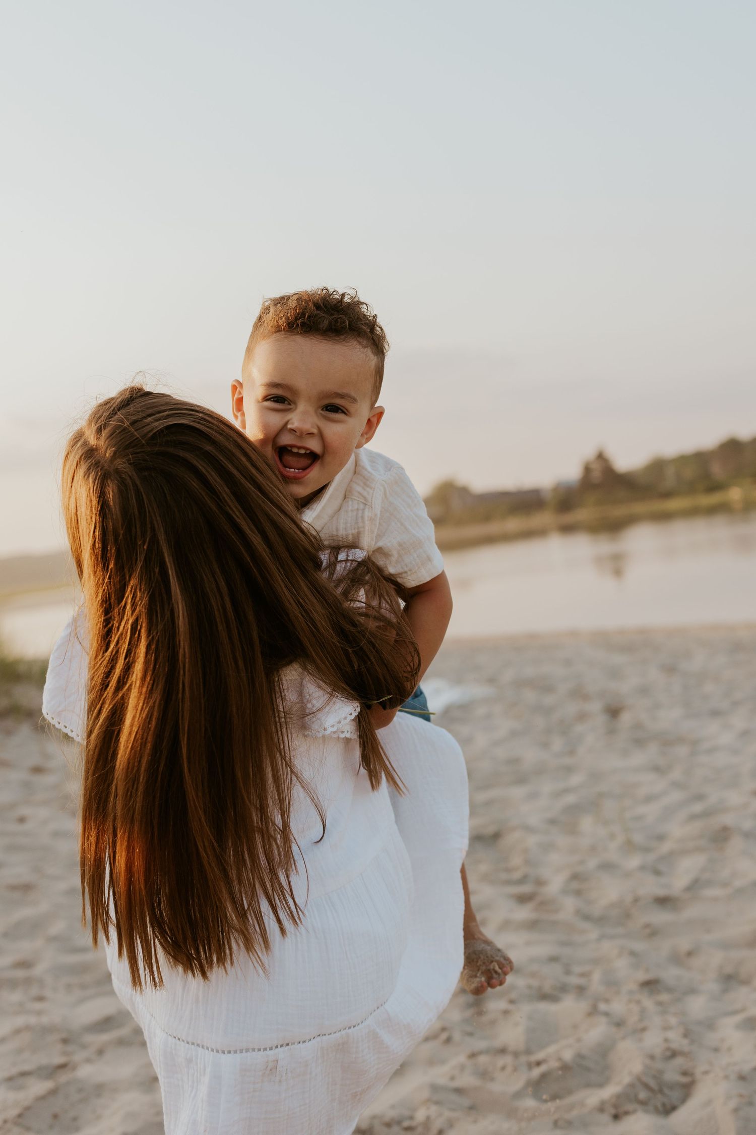 Mother lifting laughing baby boy on sandy beach at sunset with water in background.