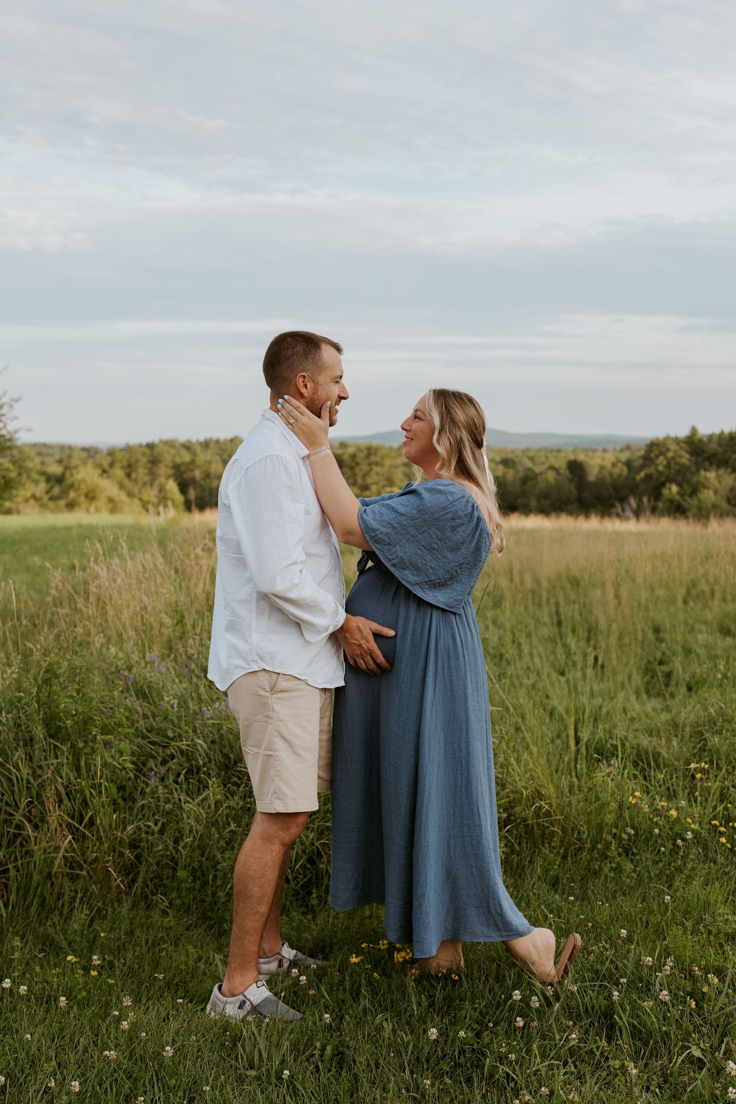 Pregnant woman in blue dress and man in white shirt embrace romantically in lush green meadow.
