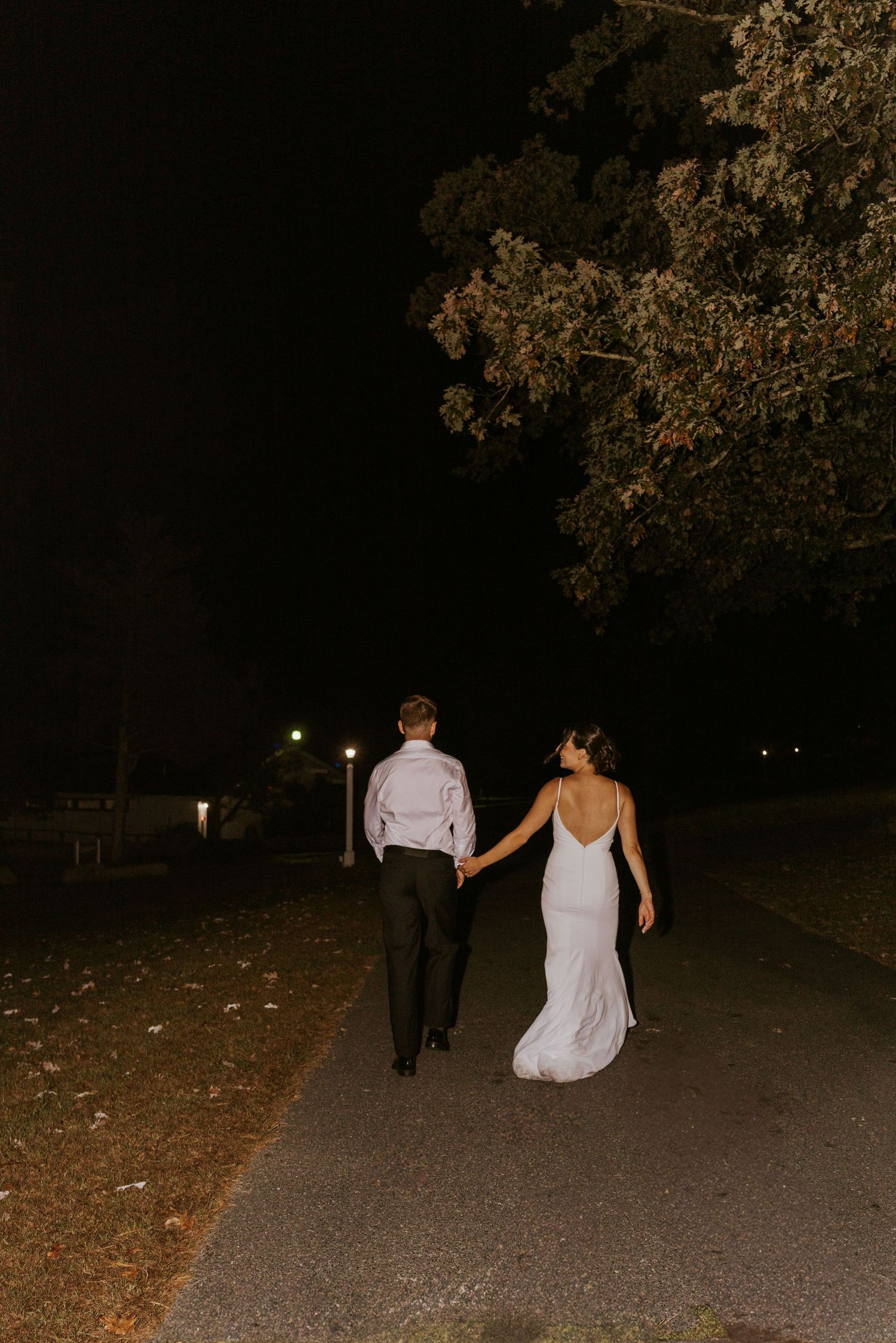 Bride in backless white gown and groom walk hand in hand down a dark tree-lined path at night.