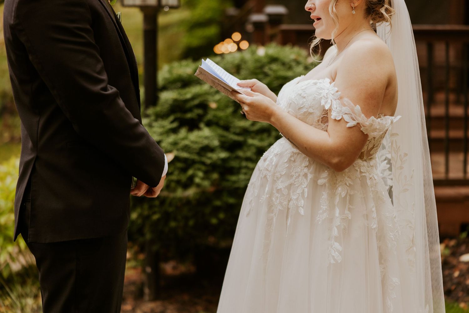 Bride in off-shoulder lace gown reads personal vows from a card during outdoor wedding first look.