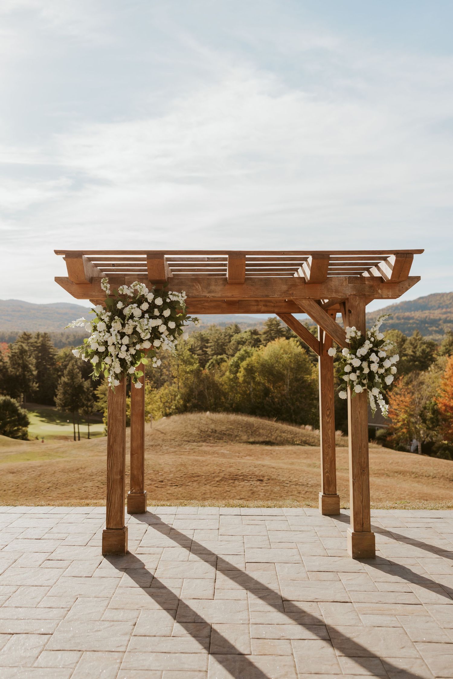 Rustic wooden pergola ceremony arch adorned with white floral arrangements overlooking autumn landscape.