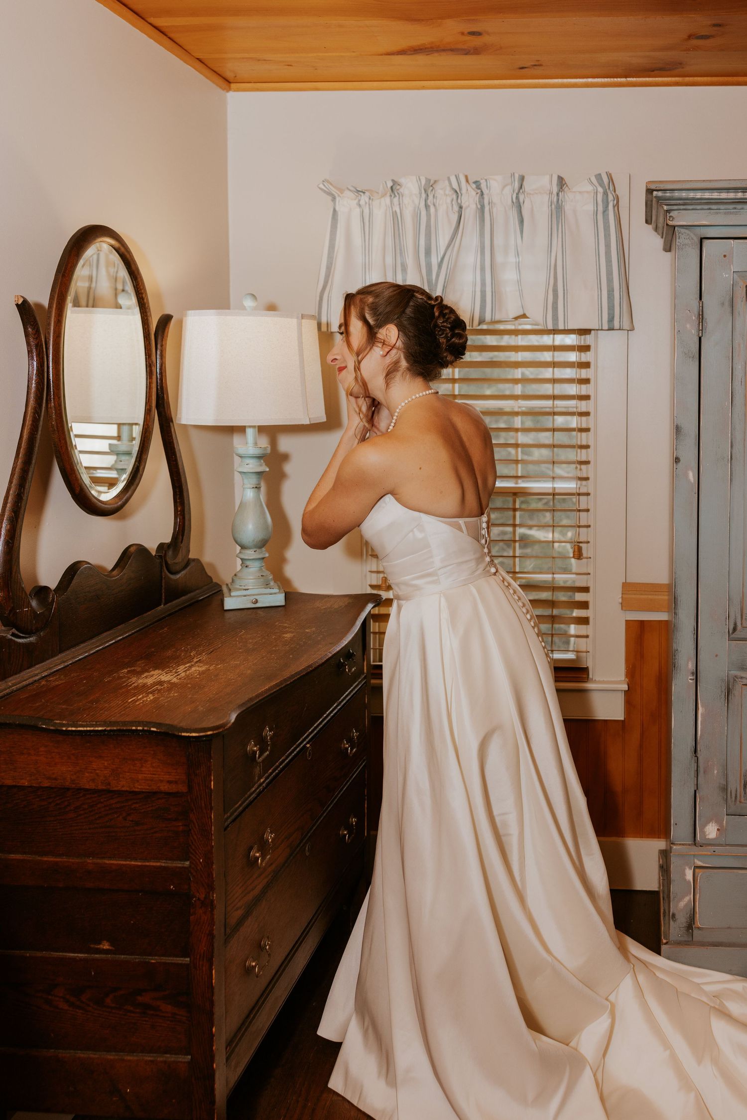 Bride in a strapless white satin gown putting on earrings beside a vintage wooden dresser with mirror.
