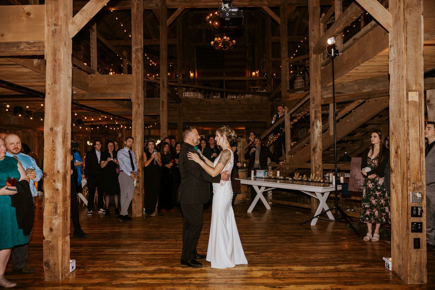 Bride and groom share first dance in rustic barn wedding venue with string lights and guests watching.