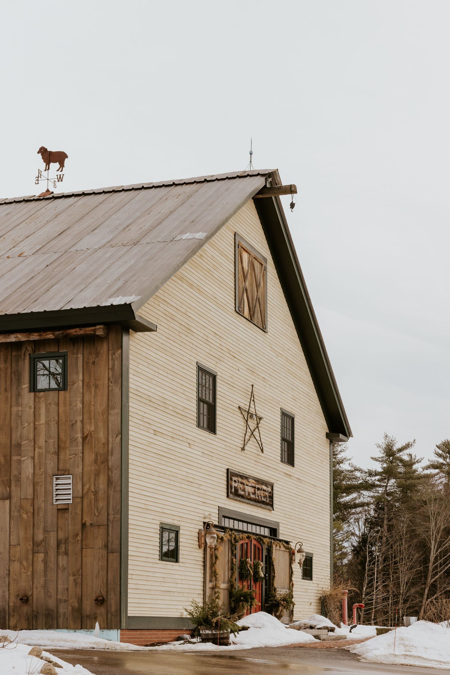 Rustic white barn with metal roof, star decor, and holiday wreaths surrounded by snow-dusted grounds in winter.