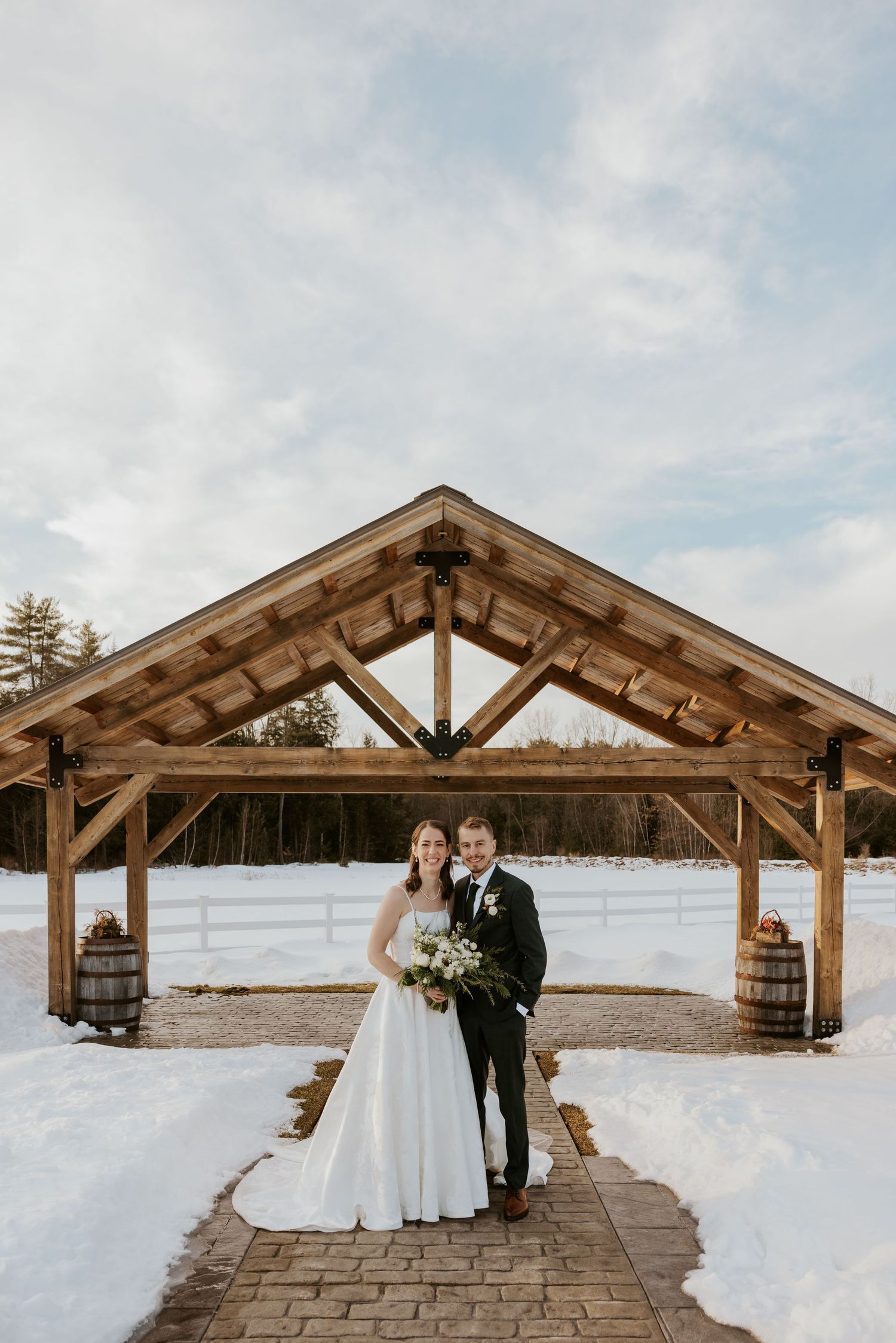 A newlywed couple poses under a rustic wooden pavilion on a snowy winter day during their wedding celebration.