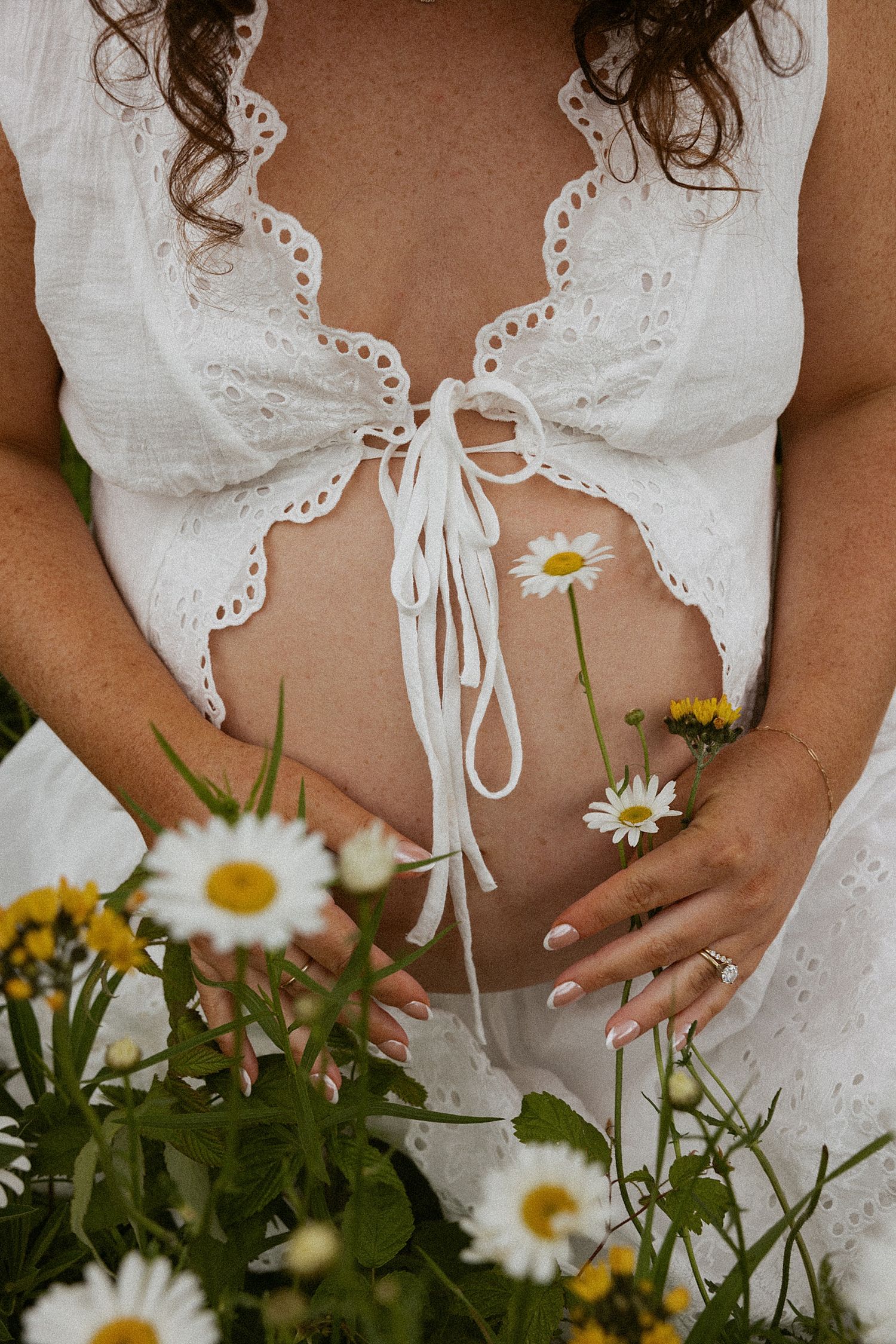 Pregnant woman in white lace crop top holding baby bump surrounded by white and yellow daisies outdoors in New Hampshire