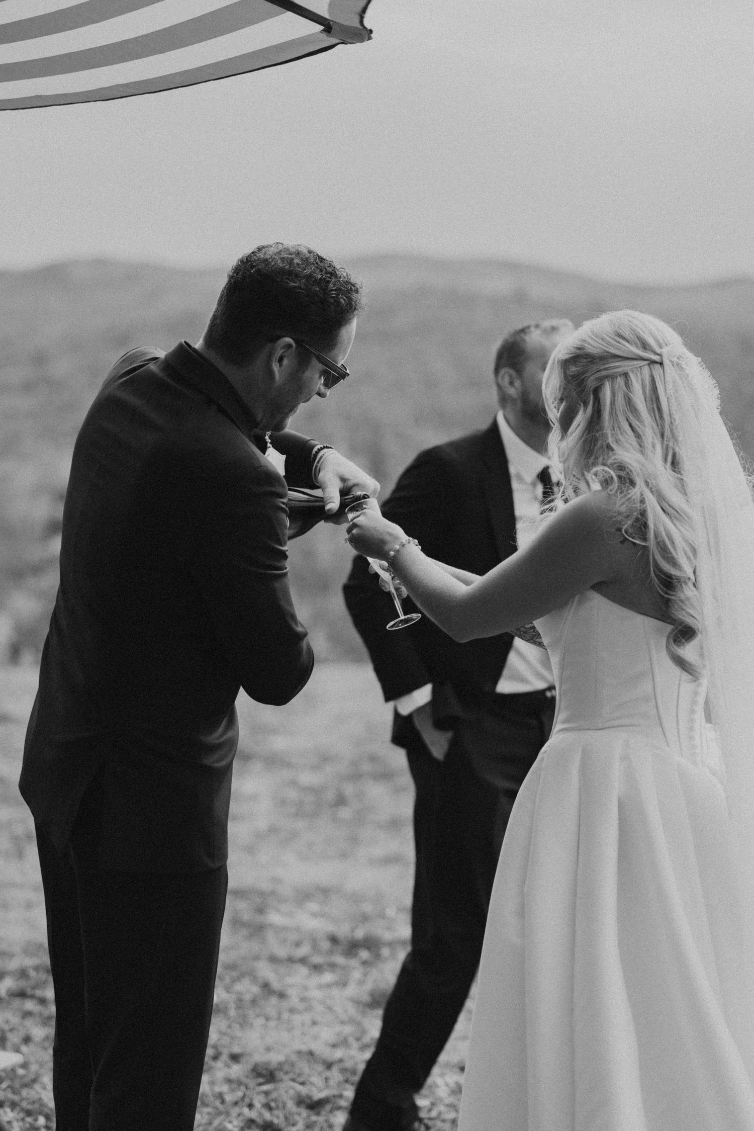 Bride and groom clinking champagne glasses outdoors with mountain backdrop in black and white.