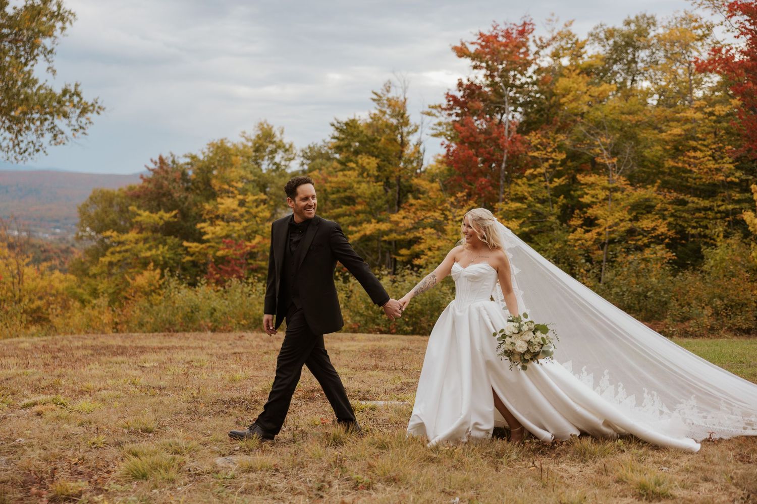 Bride and groom walk hand-in-hand through a field with vibrant autumn foliage in the background.