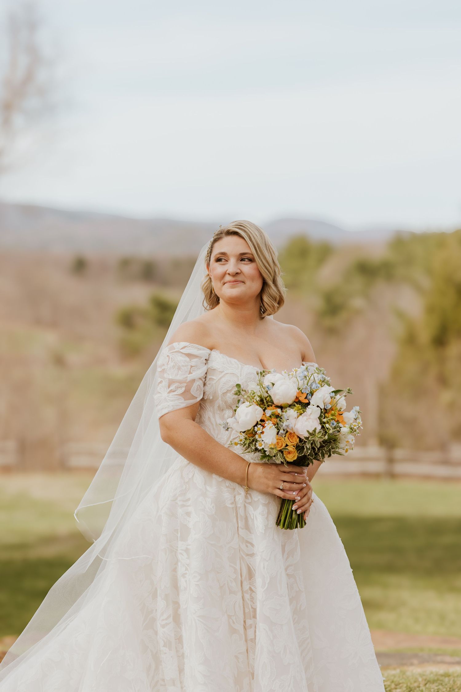 Bride in off-shoulder lace gown holding white and orange bouquet with mountain landscape background.