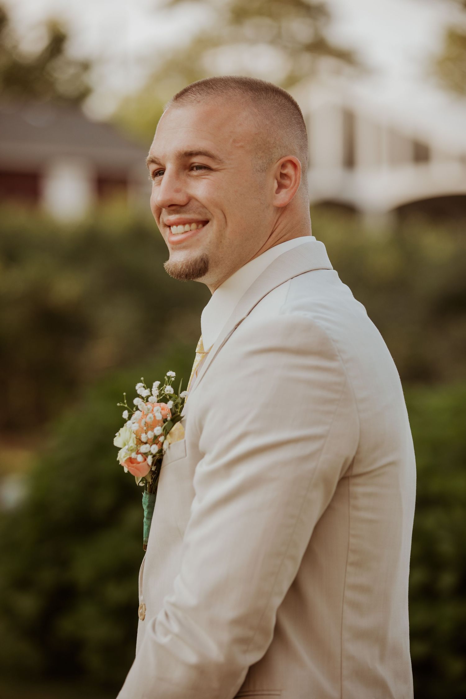 Smiling groom in beige suit with floral boutonniere posing outdoors at wedding ceremony with greenery background.