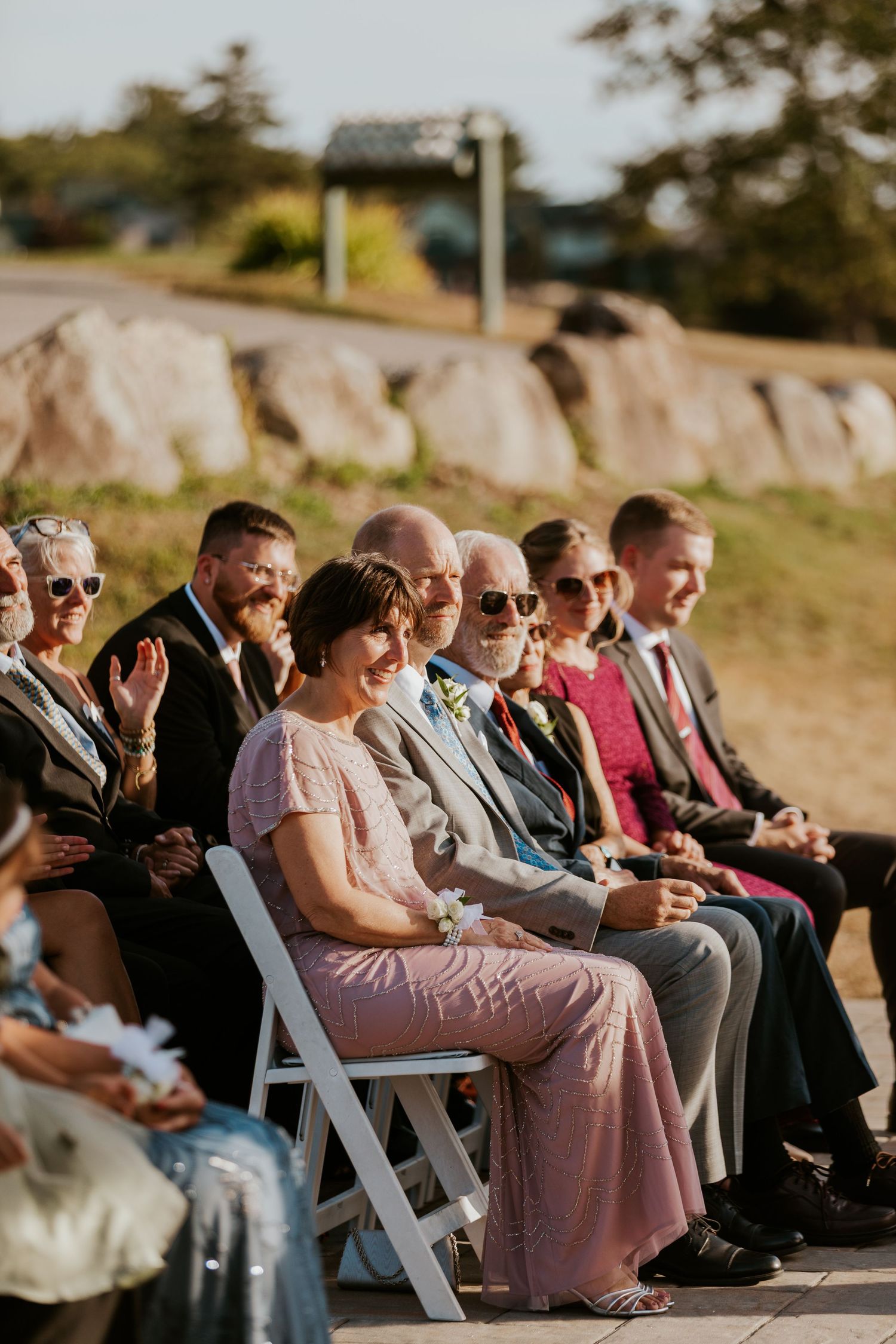 Wedding guests seated outdoors in sunlight smiling during open-air ceremony on a sunny day.