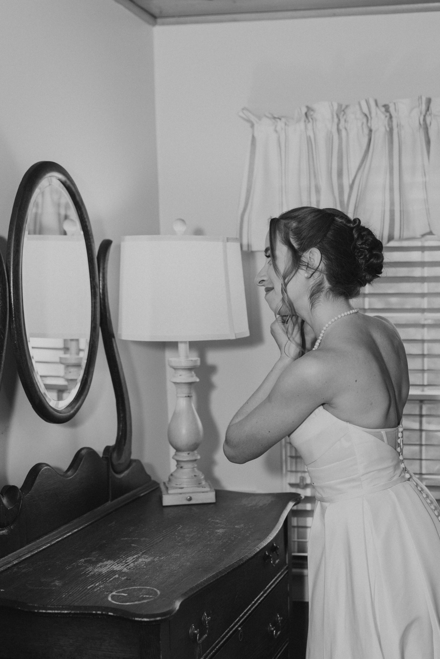 Black and white photo of bride in strapless gown putting on earrings beside antique dresser and lamp.