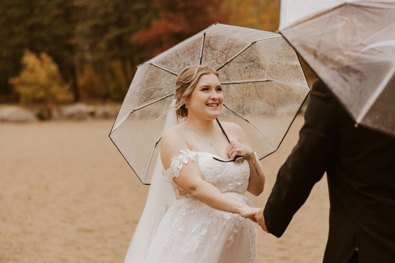 Smiling bride holds a clear umbrella on a sandy beach surrounded by vibrant fall foliage.
