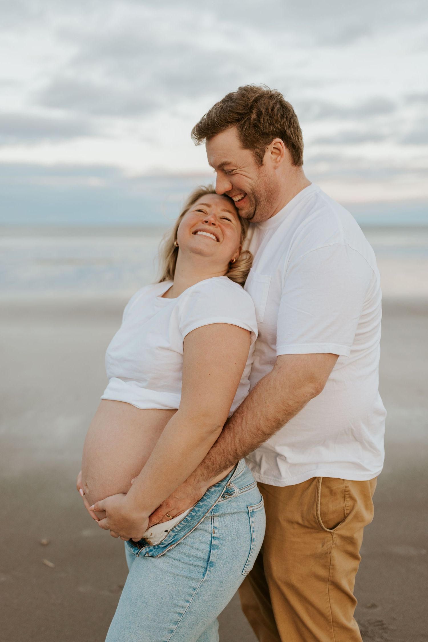 Happy expecting couple embracing on beach at sunset, both wearing white shirts.