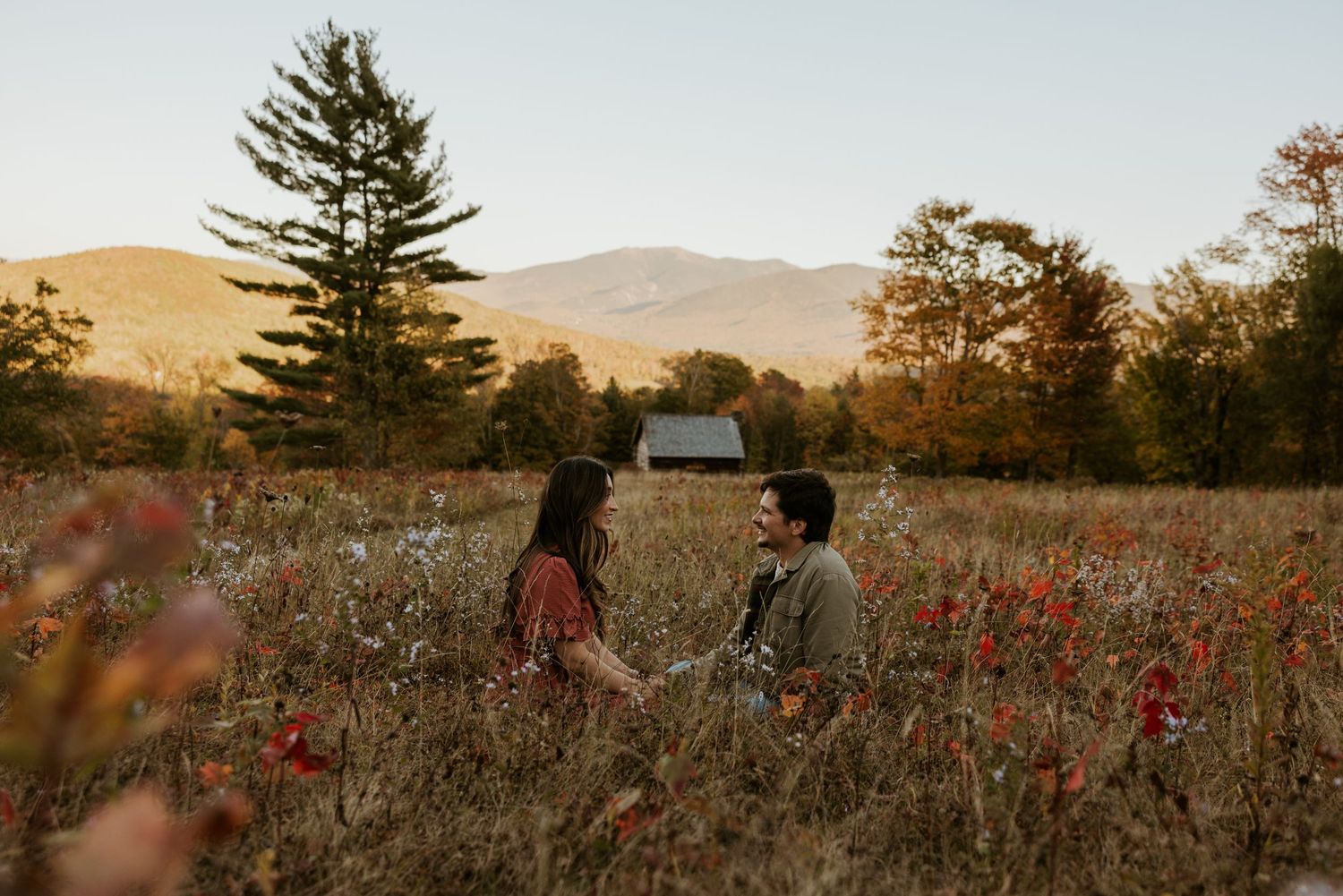 Two people holding hands seated in vibrant red wildflower field with rustic barn and mountains.