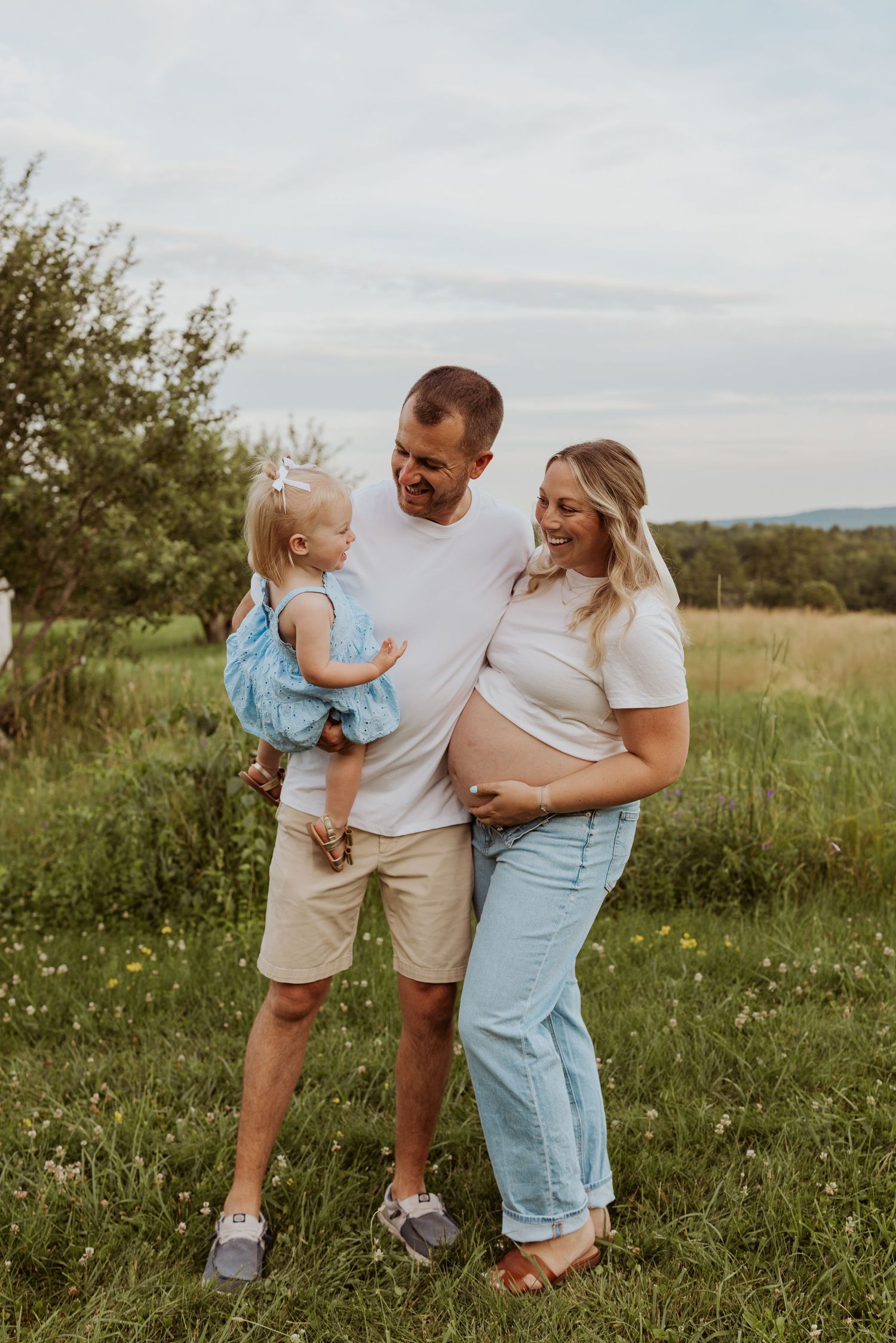 Pregnant woman in white top and jeans leans on partner holding toddler in a green meadow.