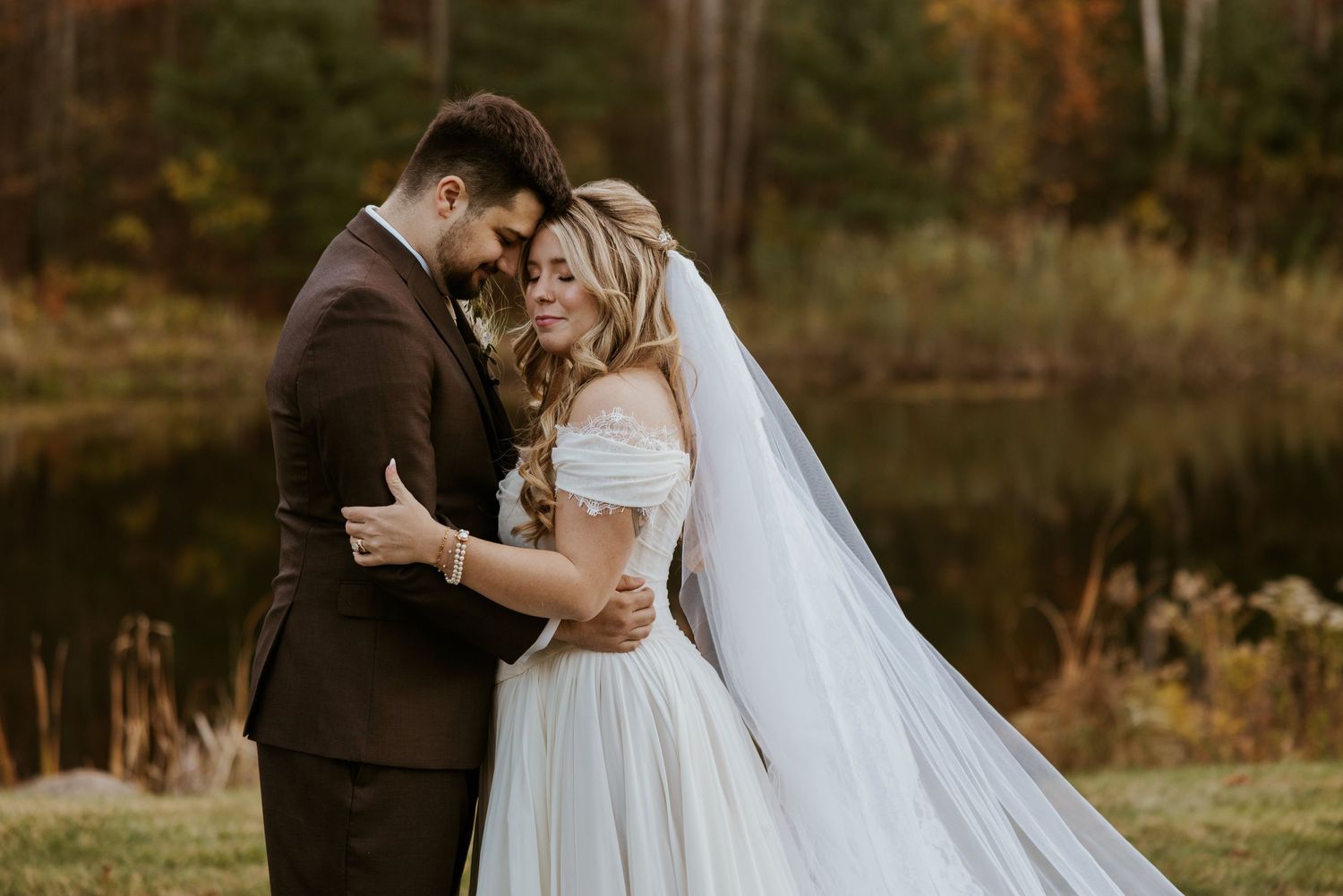 Romantic couple embraces in an off-shoulder ballgown with flowing veil beside an autumn pond.