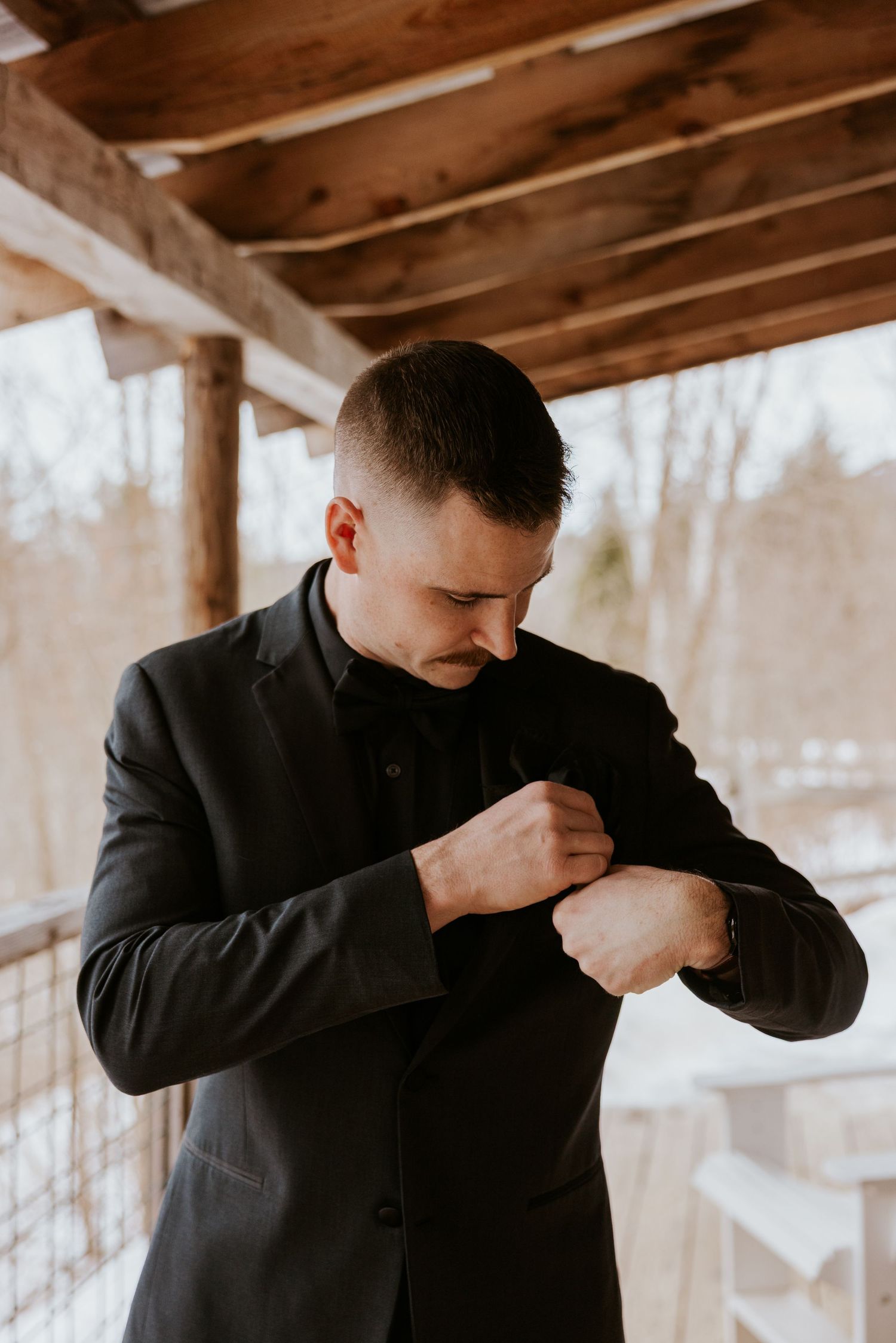 Groom in black tuxedo adjusting his cufflinks on a wooden porch with snowy winter backdrop.