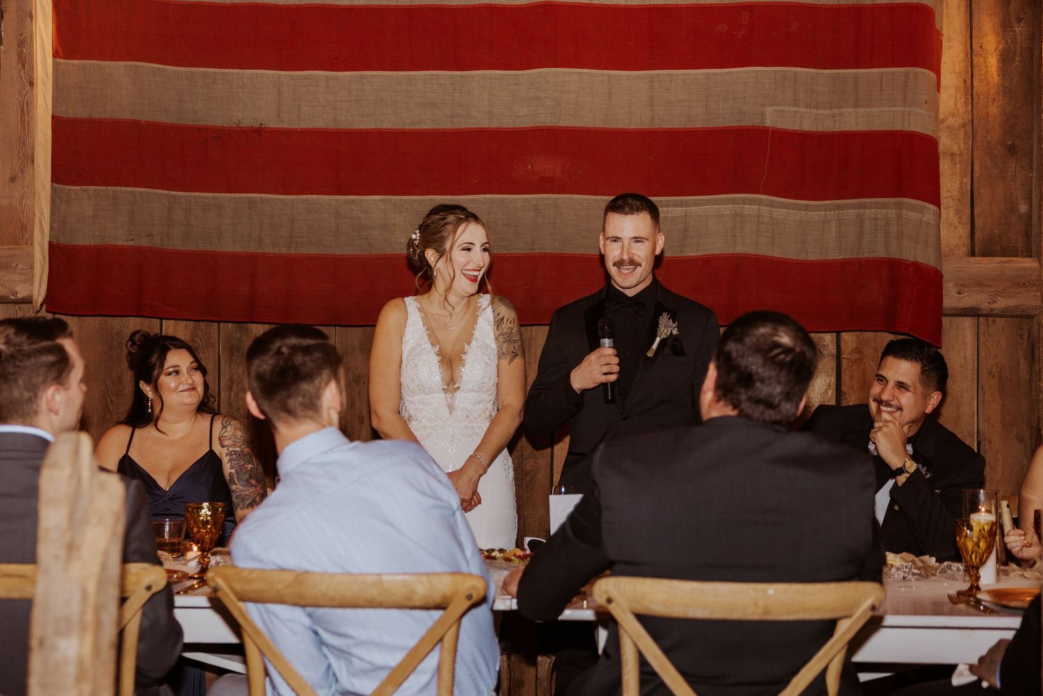 Bride and groom stand together during wedding reception toast in rustic barn venue with guests seated around table.