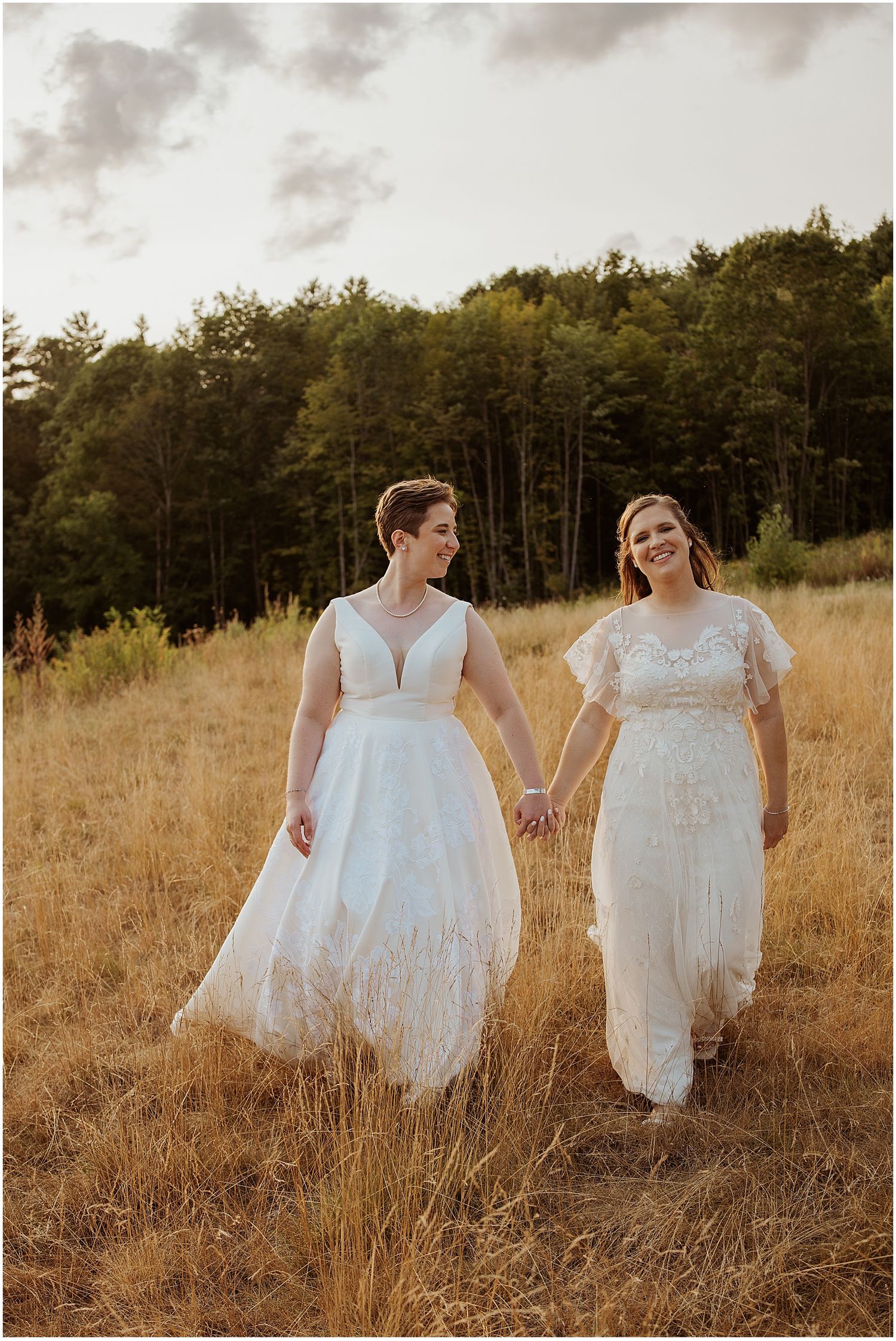 Two people in white dresses walk through a golden field at sunset holding hands and smiling.