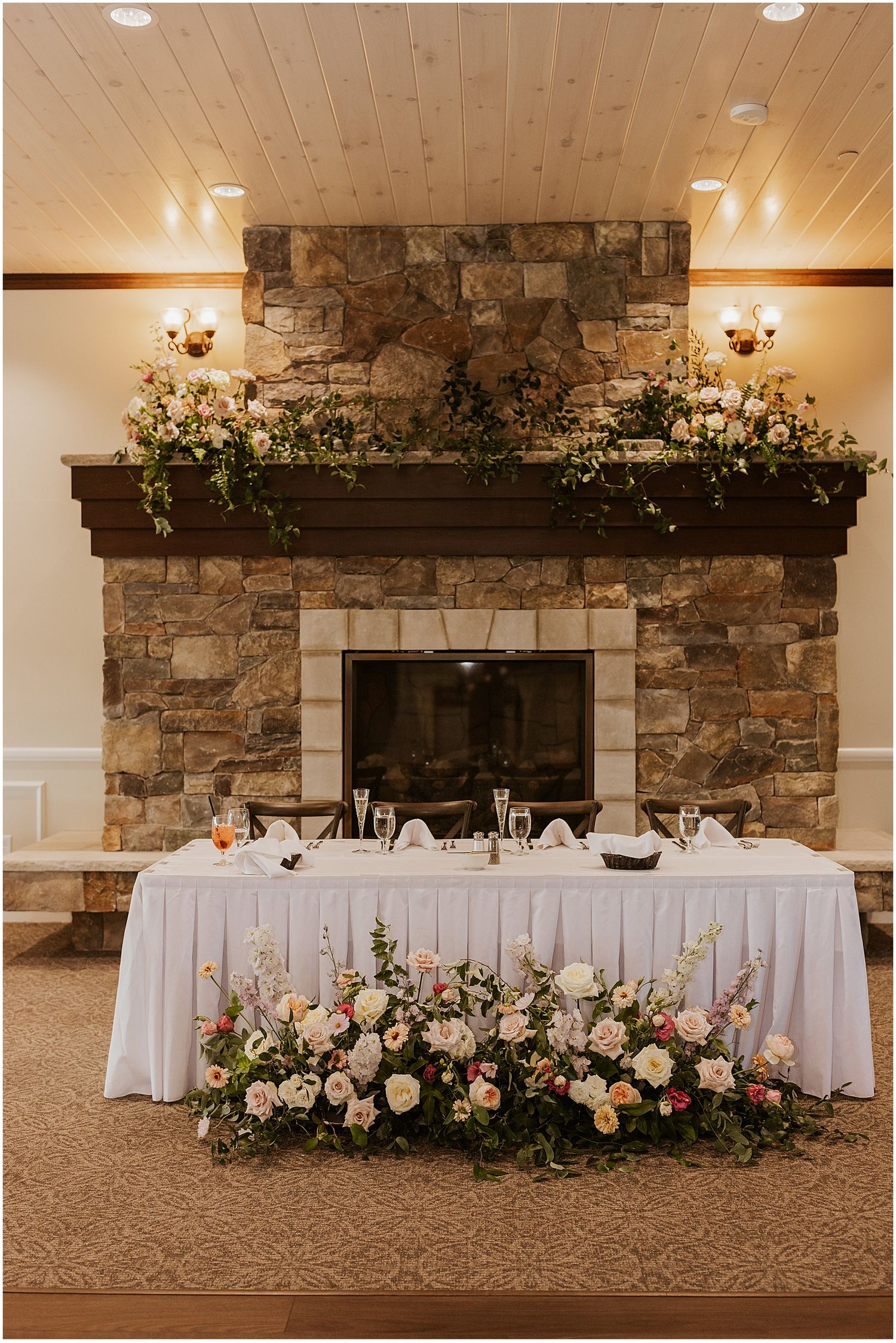 Elegant wedding reception head table decorated with lush floral arrangements and stone fireplace backdrop.