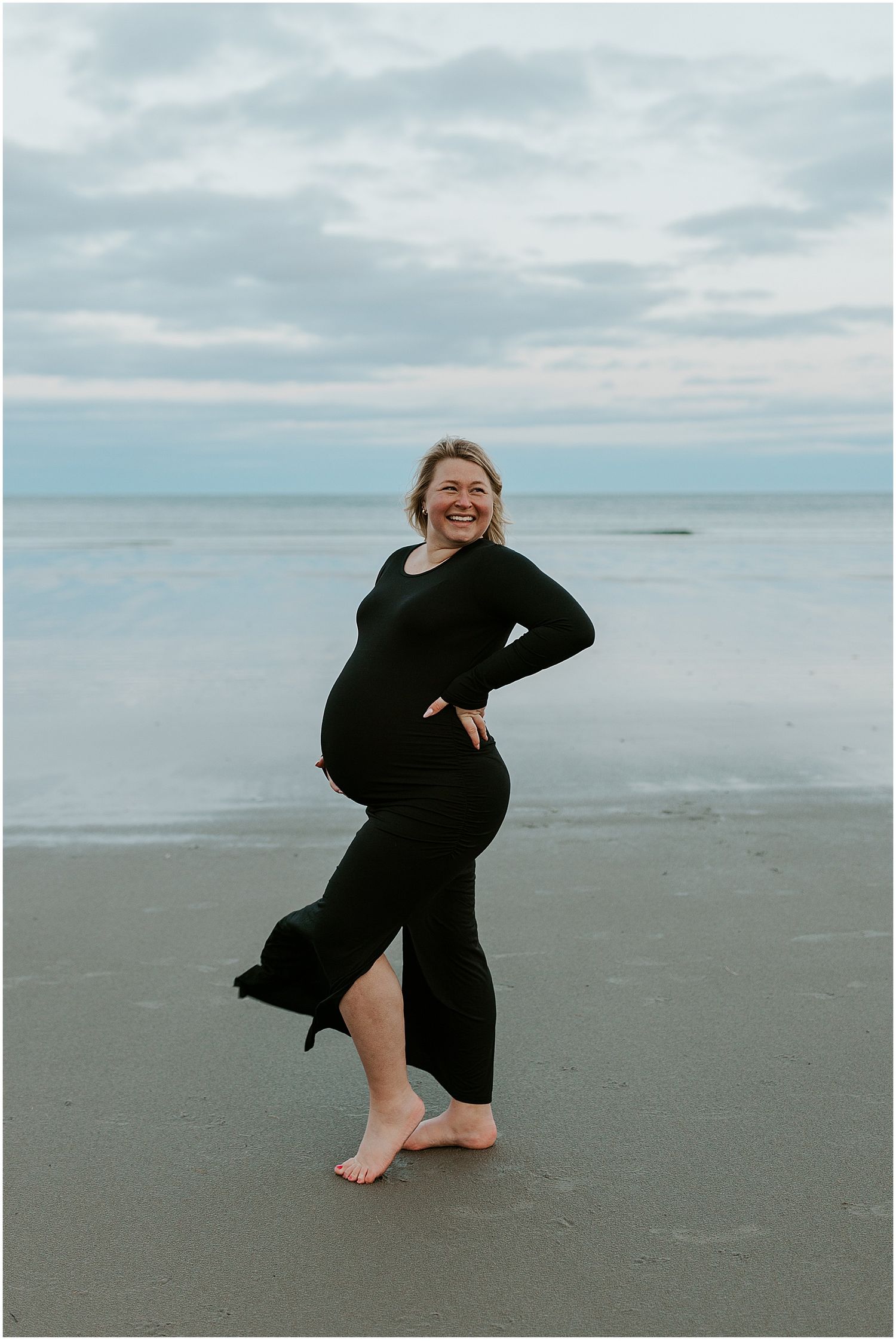 Person in black performs graceful dance moves on an empty beach under cloudy skies.