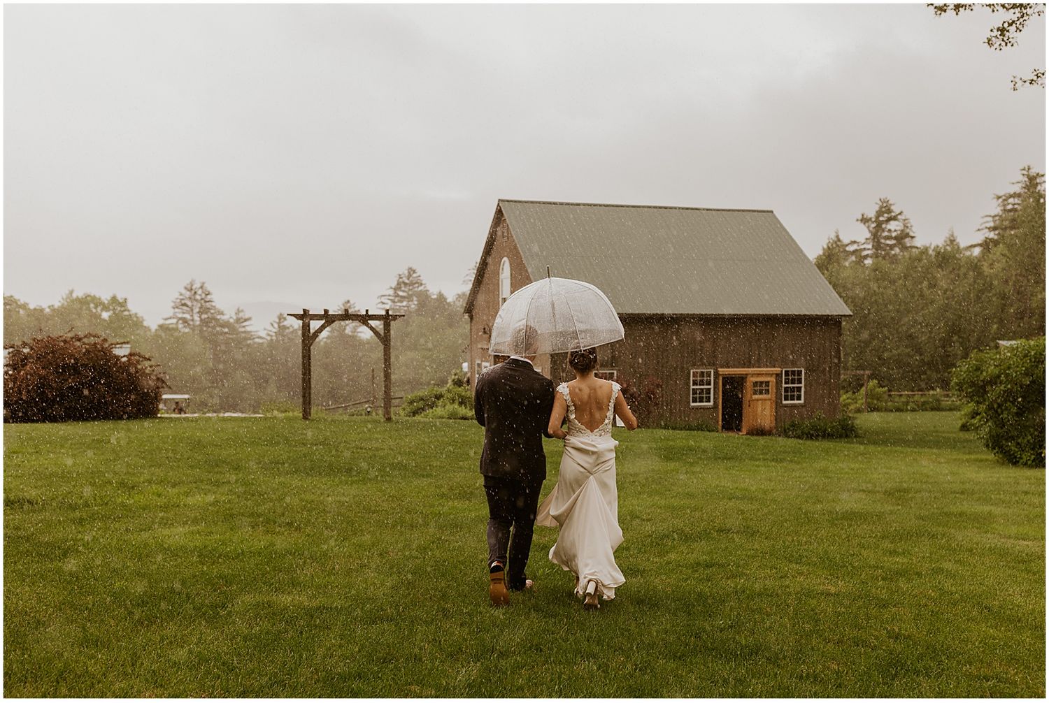 A couple walks through a rustic field with farm buildings in the distance.