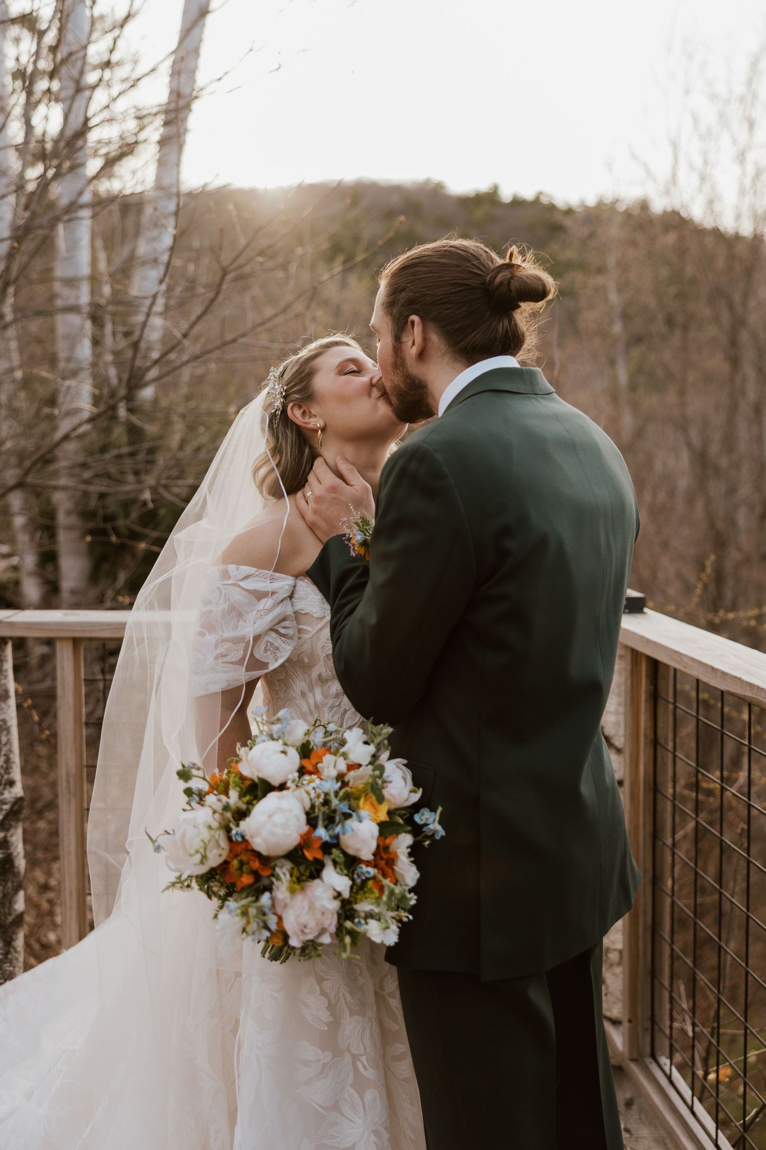 Bride and groom kissing on a wooden deck at golden hour with colorful bridal bouquet.