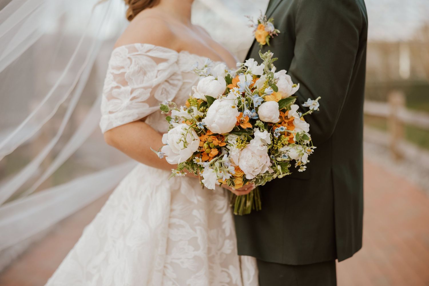 Bride and groom holding a lush wedding bouquet with white peonies and orange blooms, bride in lace off-shoulder gown.