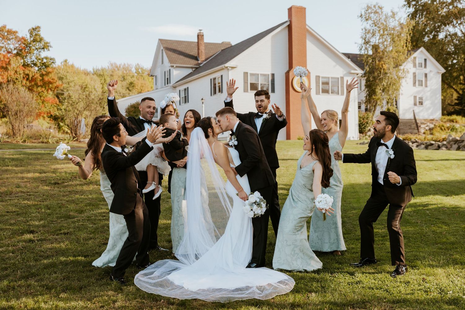 Wedding party celebrates with bride and groom kissing surrounded by bridesmaids and groomsmen outdoors.