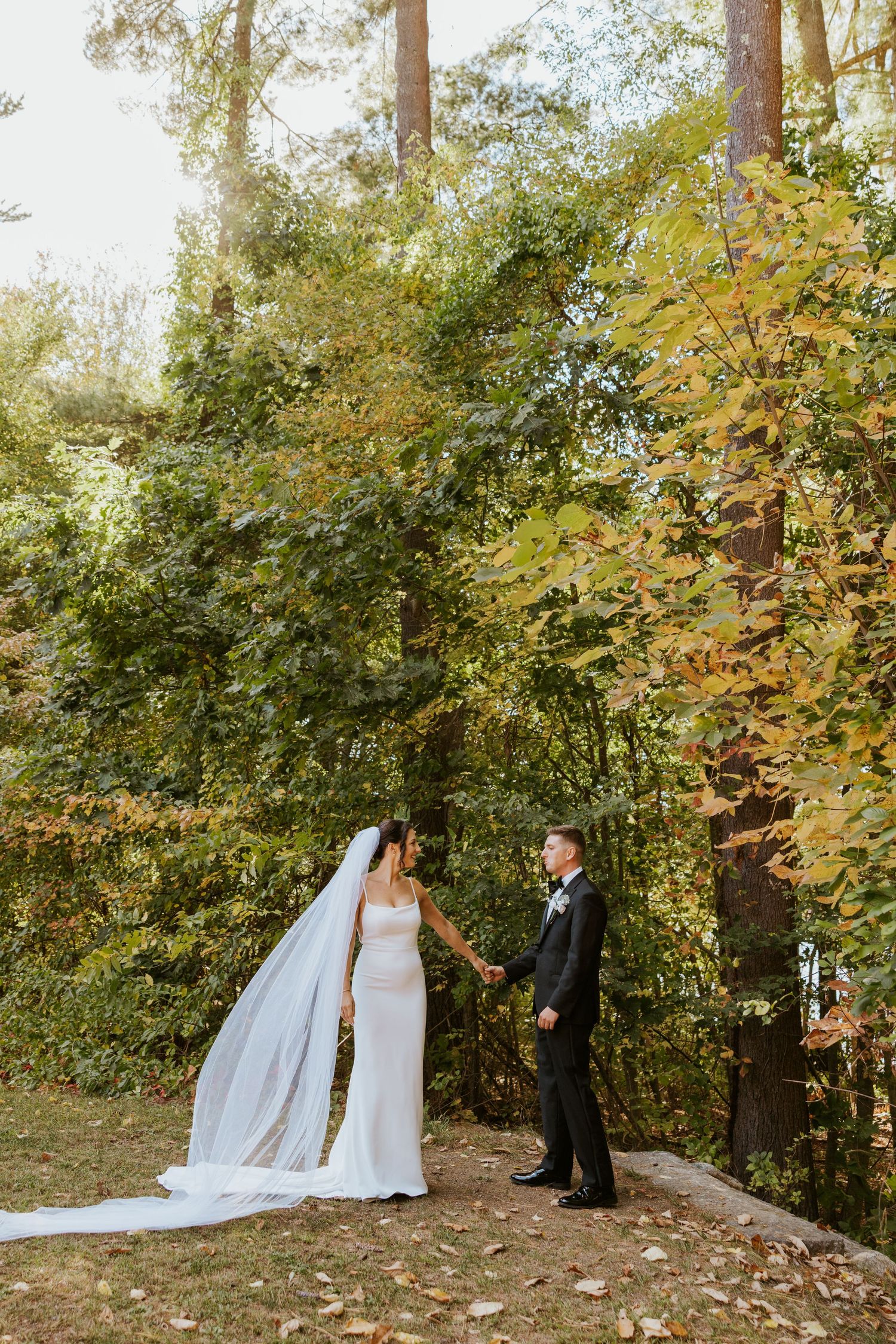 Bride and groom holding hands outdoors surrounded by lush green and golden autumn trees.