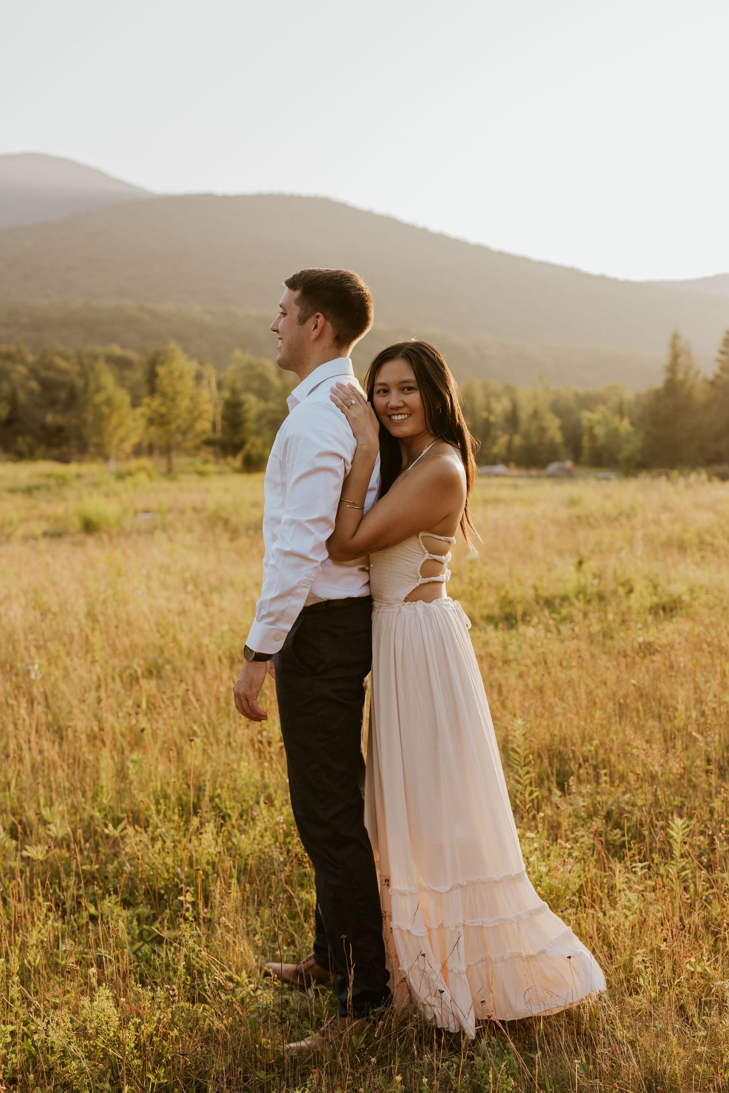 Bride in blush gown embraces groom from behind smiling in golden mountain meadow.