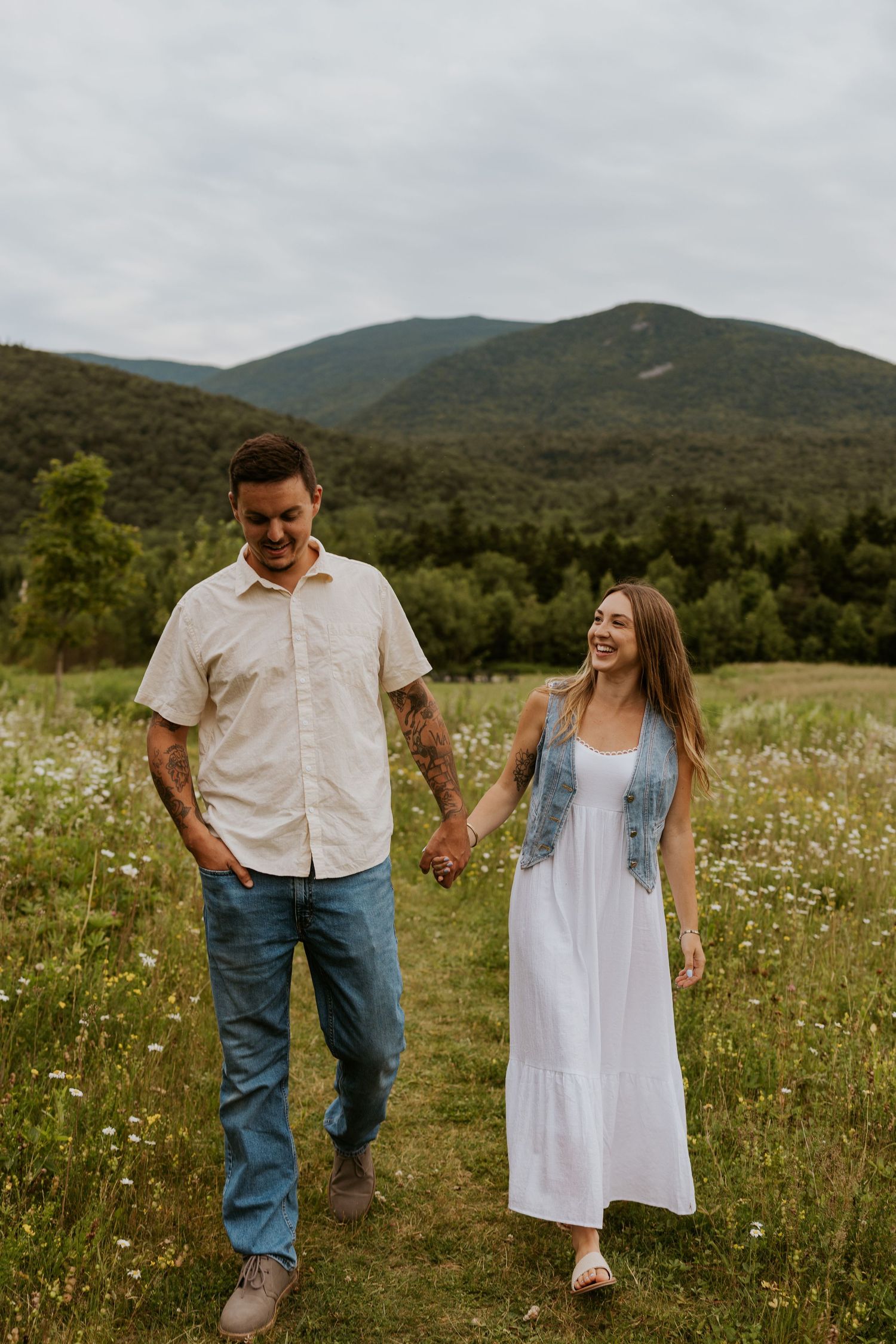 Smiling couple strolling hand in hand through daisy-filled field with green mountain backdrop.