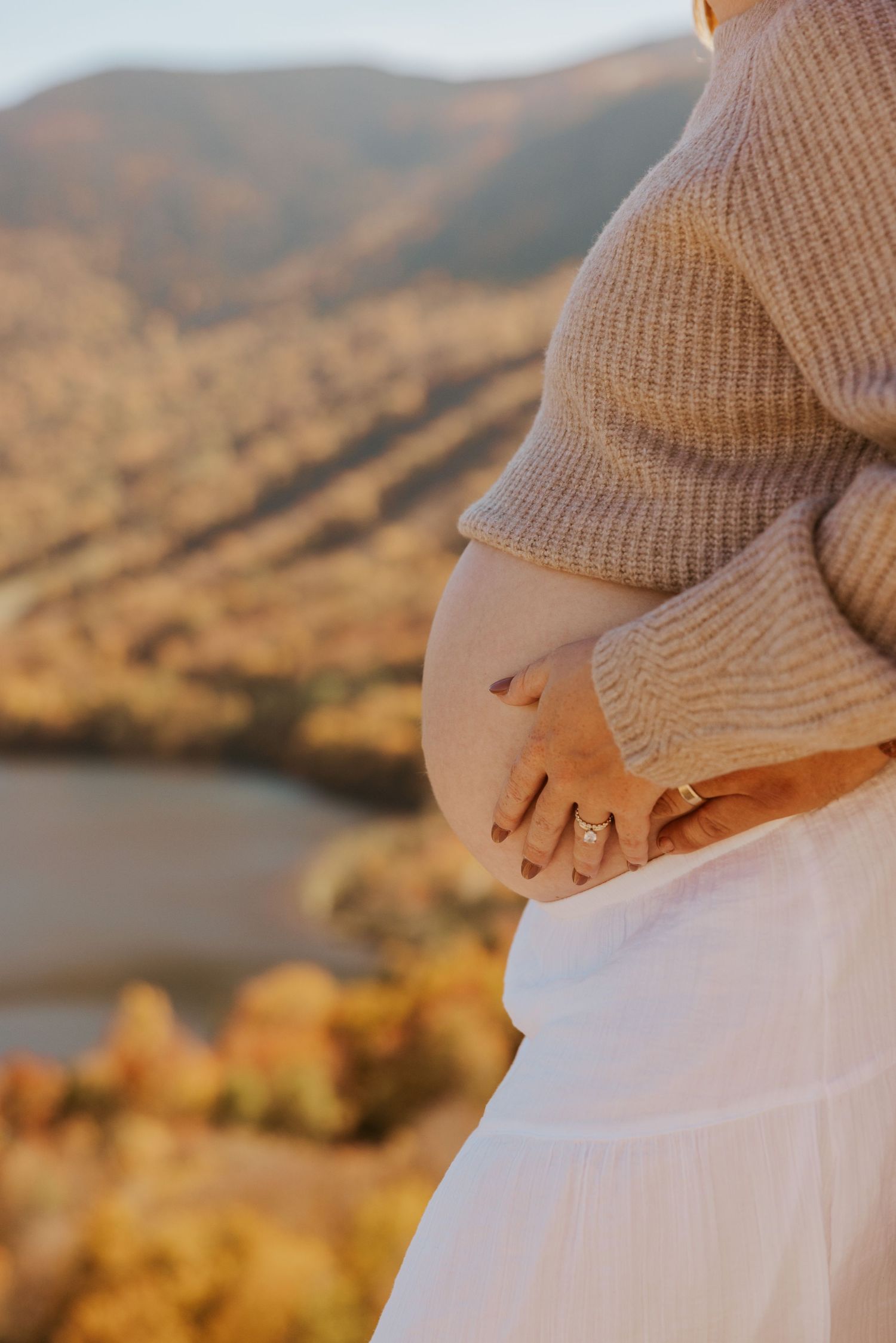 Pregnant woman in beige sweater and white skirt cradling baby bump with mountain autumn landscape background.