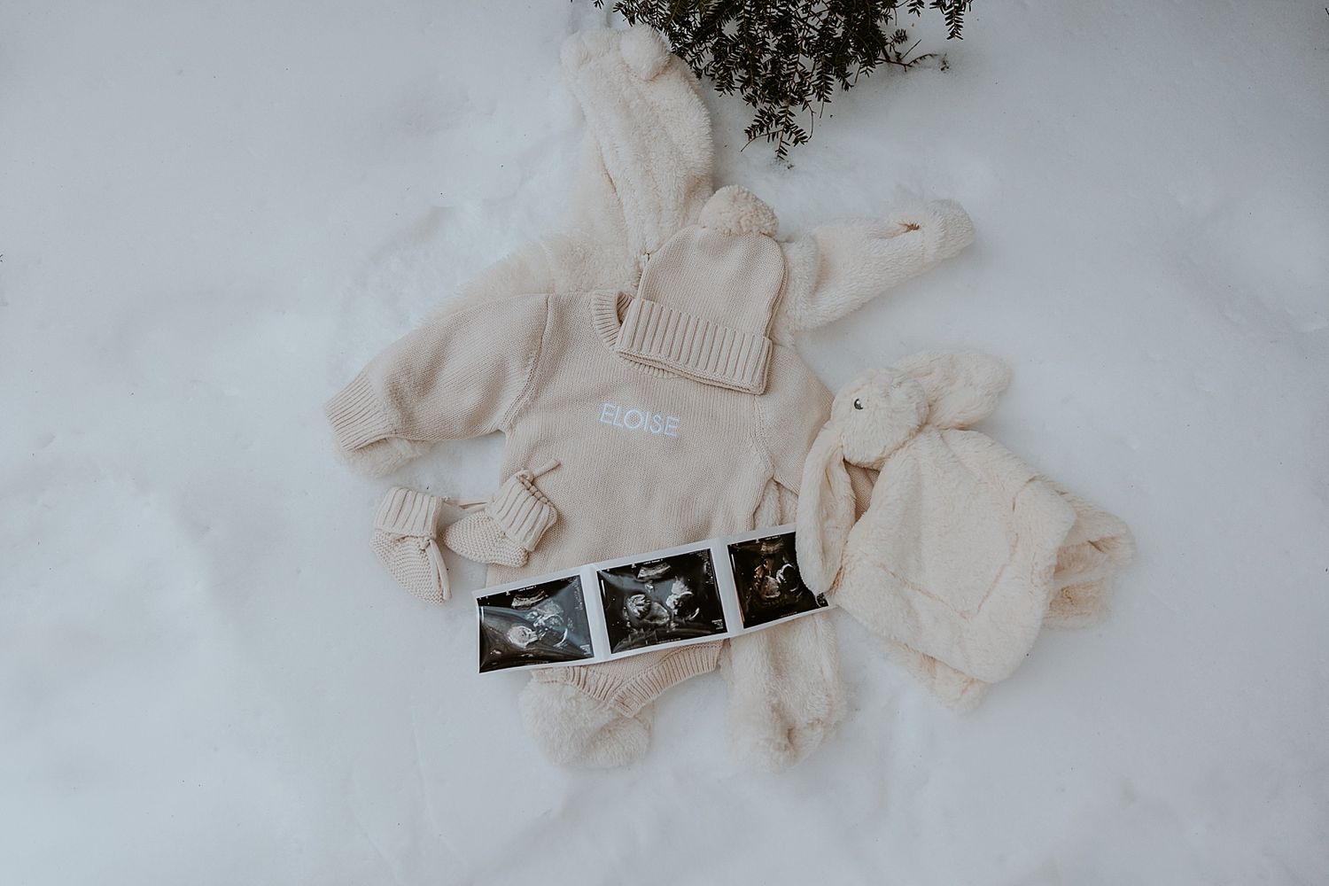 Baby announcement flat lay with ultrasound photos, knit hat, booties, sweater, and stuffed bunny on white snow in New Hampshire