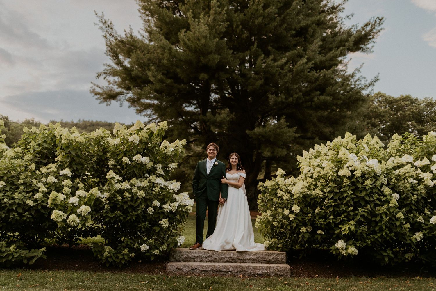 Newlyweds pose together on stone steps surrounded by white hydrangea bushes in a garden setting.