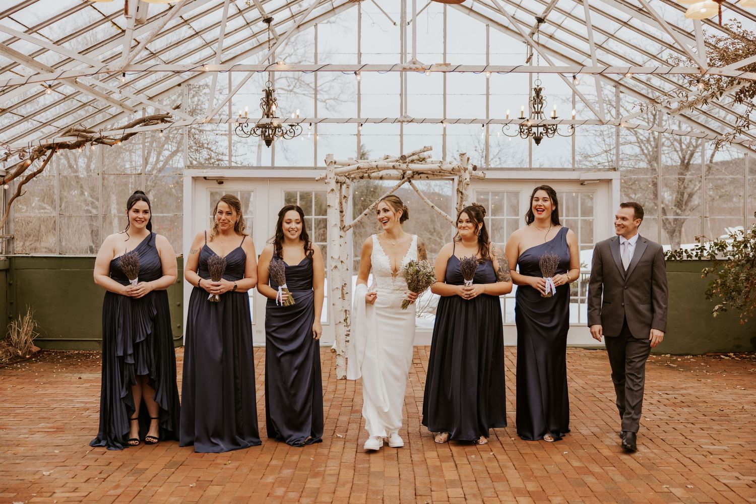 Bride with bridesmaids in navy gowns and groomsman pose in elegant greenhouse wedding venue.