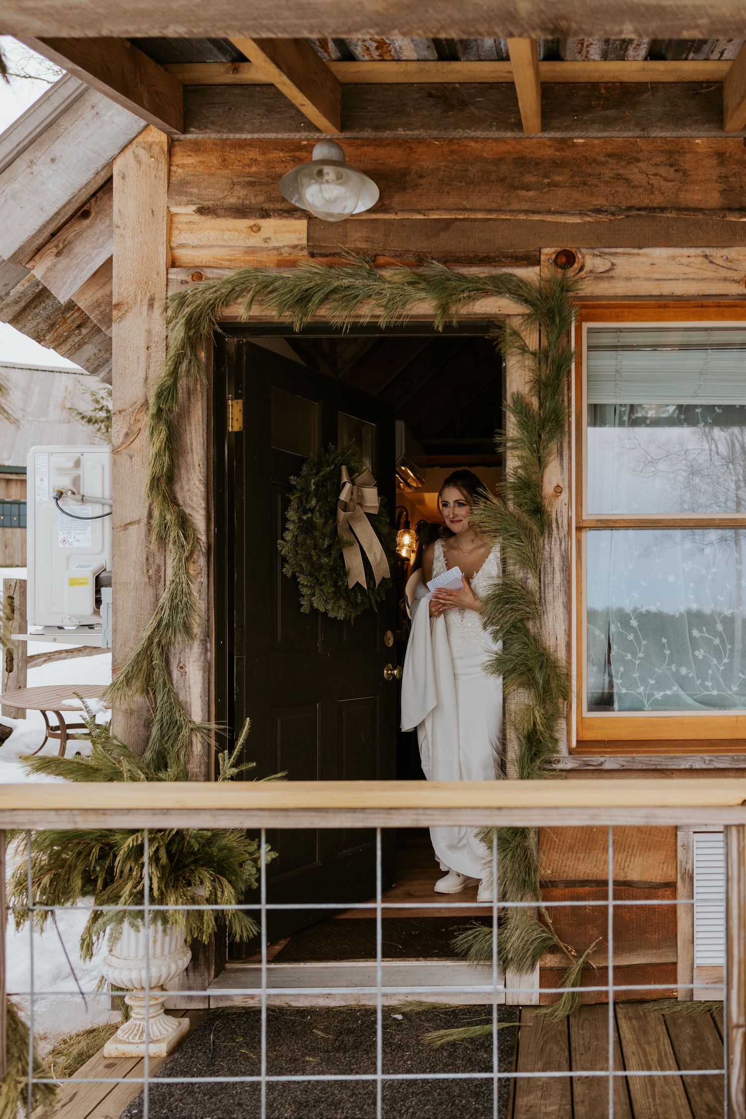 Bride in white gown standing in doorway of rustic cabin decorated with greenery.