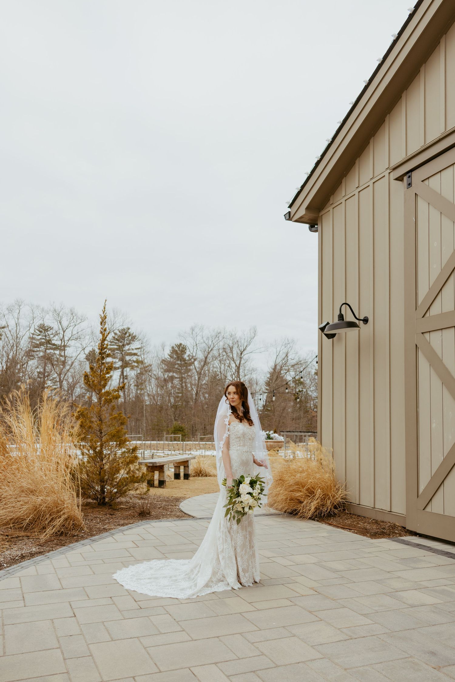 A modern barn-style venue exterior with ornate stone patio and decorative grasses on a cloudy day.