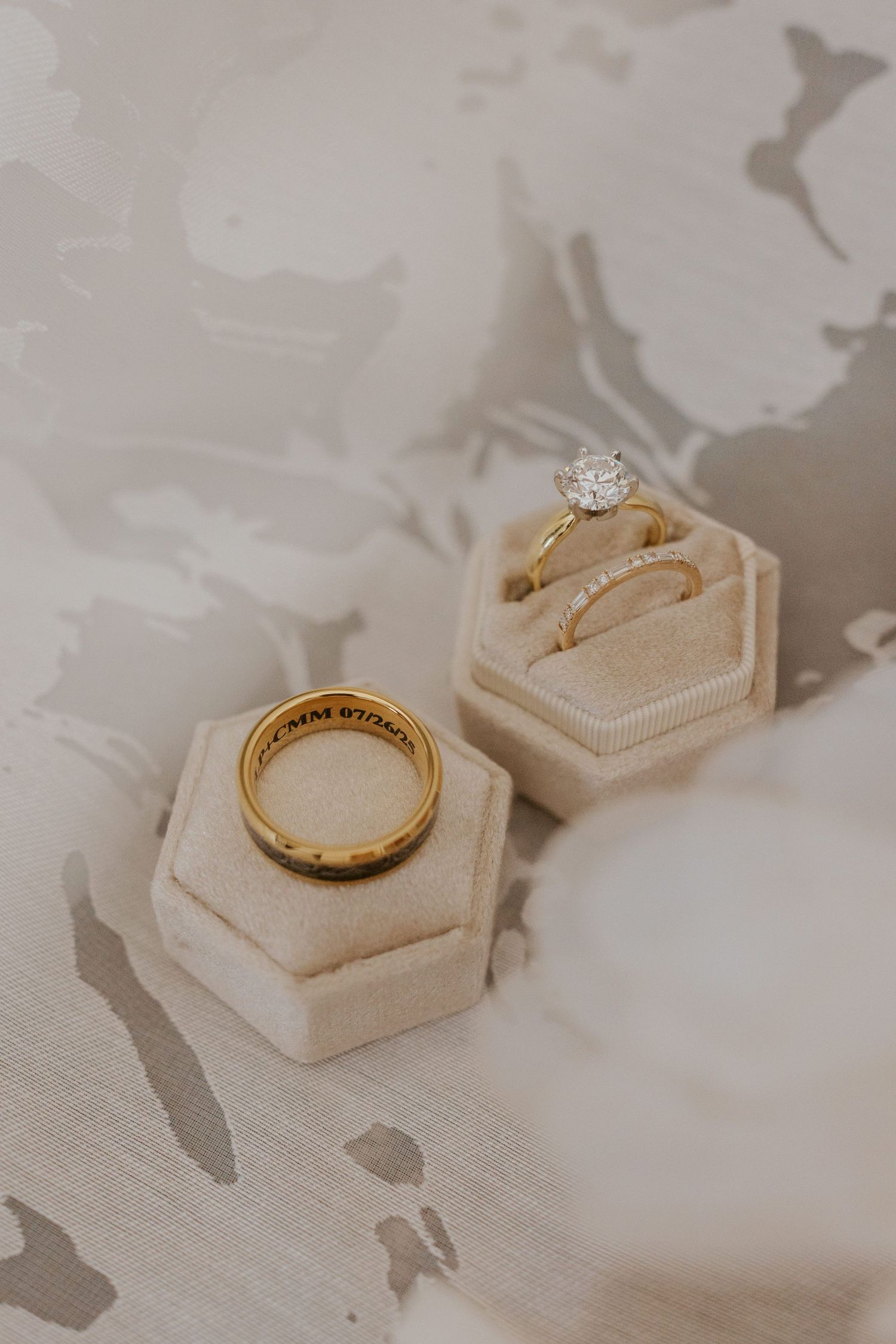 Two gold wedding rings displayed in elegant hexagonal velvet ring boxes on a floral marble surface.