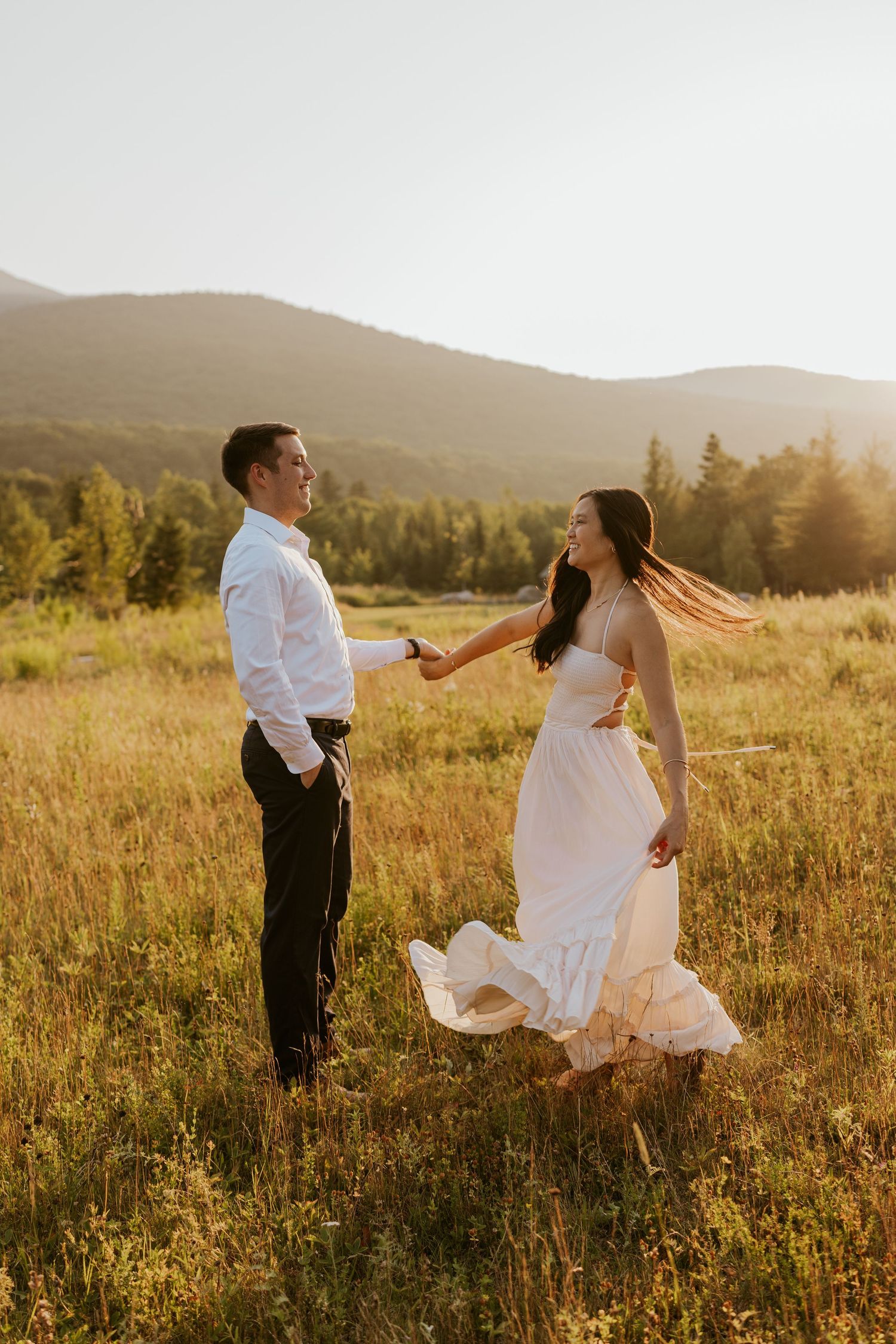 Woman in flowing white dress twirls toward partner in sunlit mountain meadow.