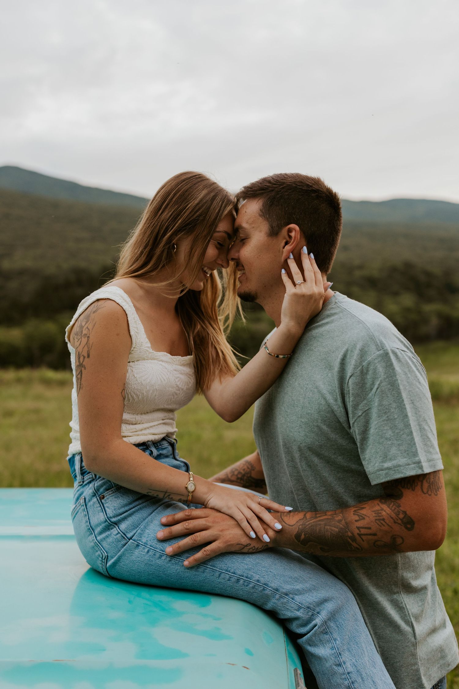Intimate couple nearly kissing on vintage turquoise car hood with mountain landscape in background.