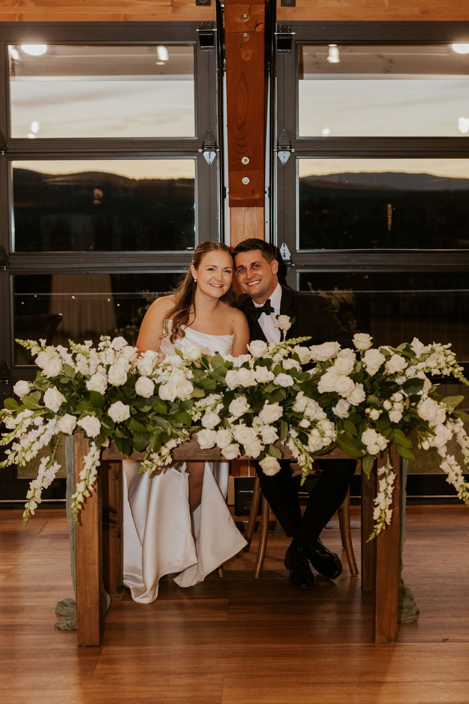 Bride and groom seated at sweetheart table adorned with lush white floral arrangements.