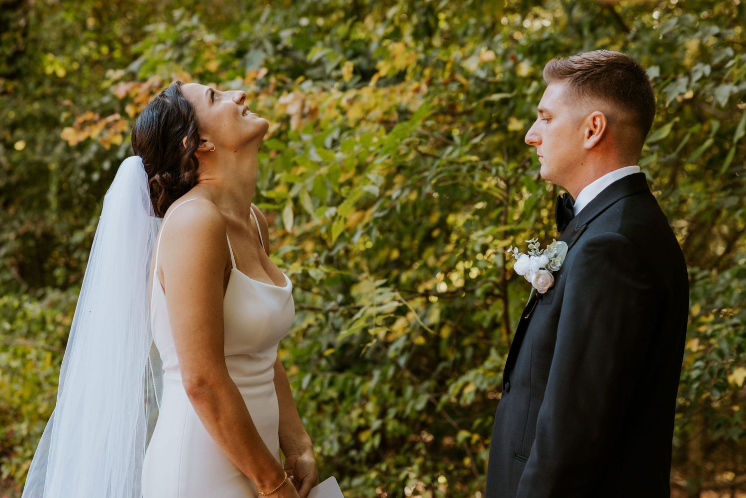 Bride laughing joyfully while holding grooms hands during outdoor wedding portraits in autumn forest.