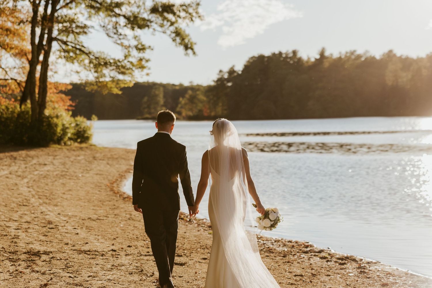 Bride and groom walk hand in hand along a sunlit sandy lakeside path in autumn.
