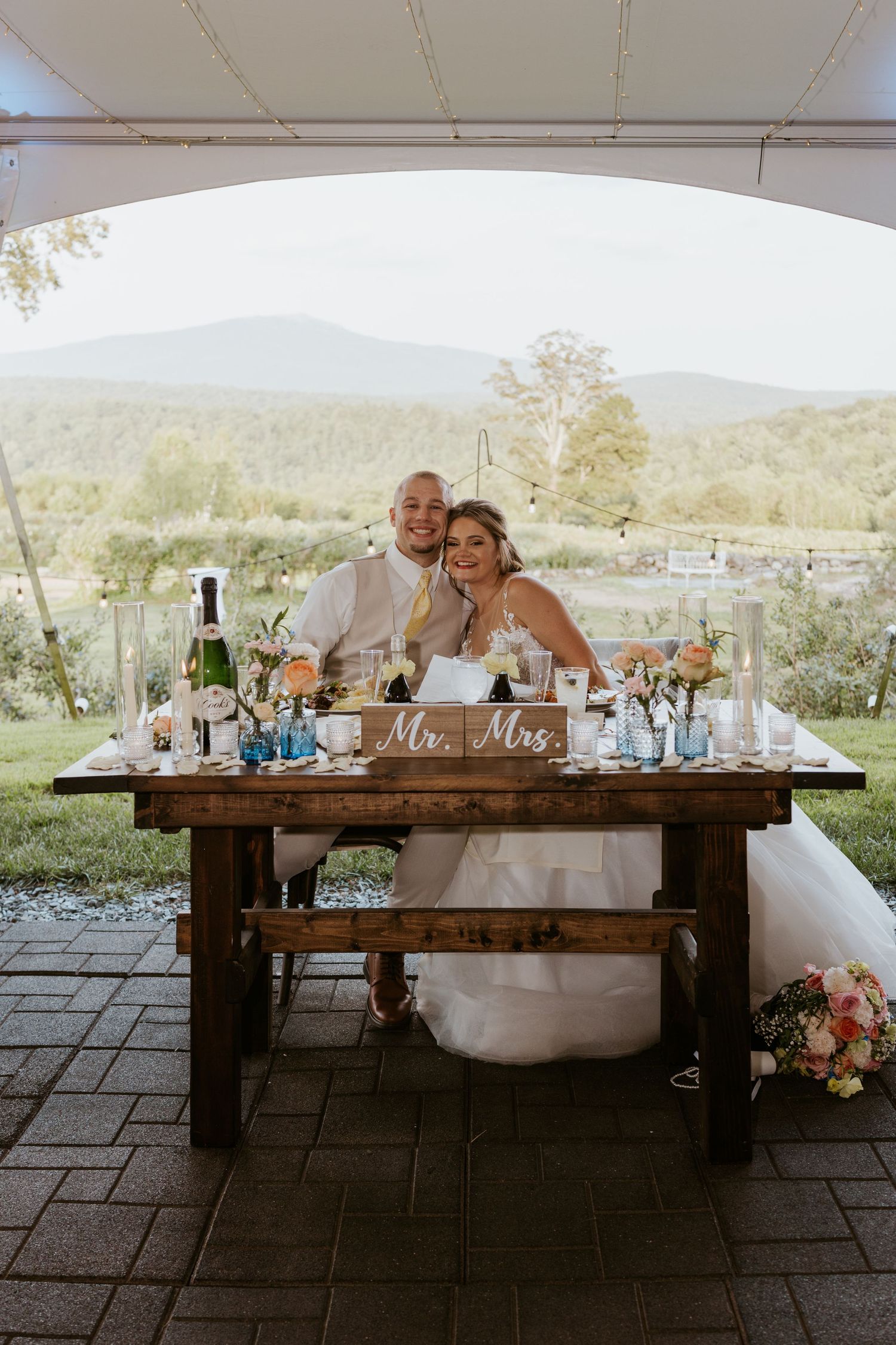 Newlyweds sit at rustic wooden sweetheart table with Mr. and Mrs. sign and mountain views behind them.