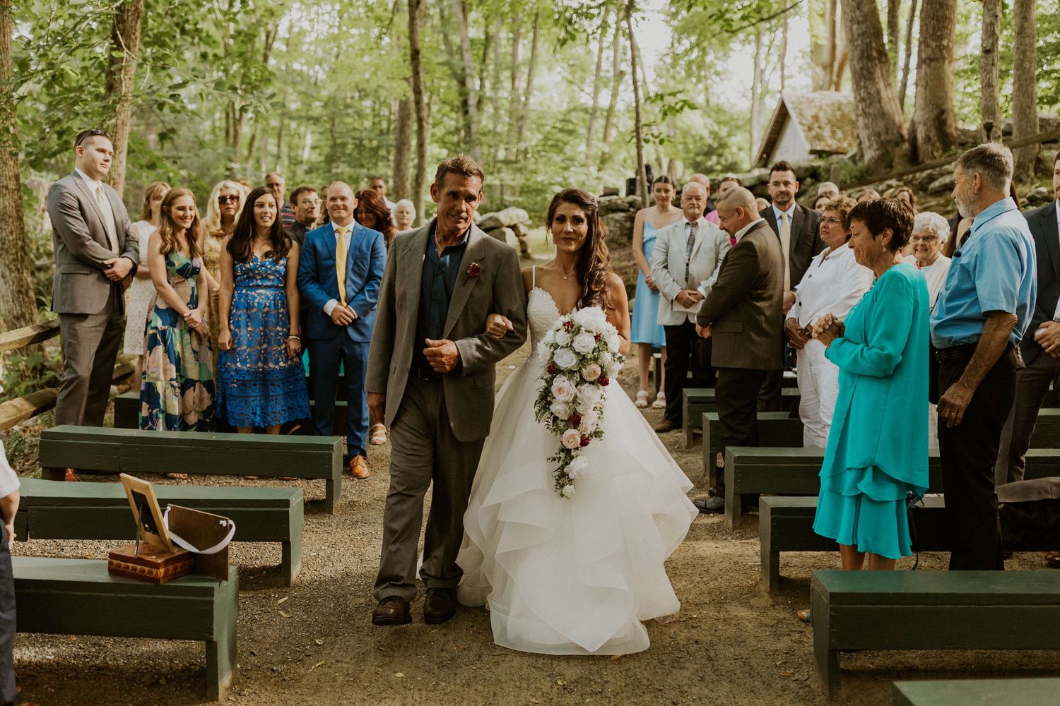 Outdoor forest wedding ceremony with guests gathered on wooden benches surrounded by trees.