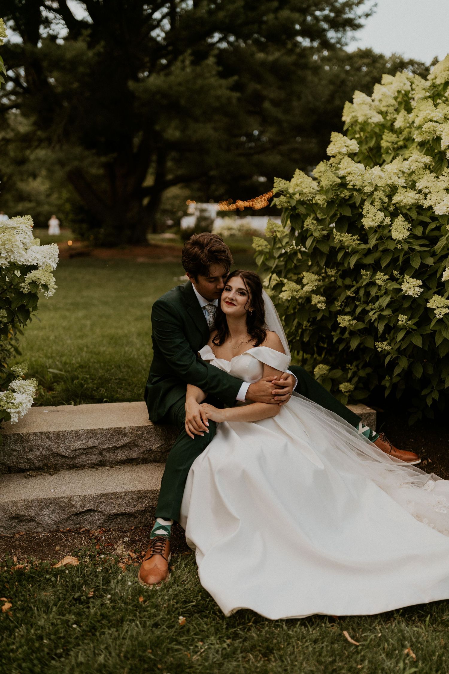 A wedding couple shares an intimate moment on stone steps surrounded by white hydrangea bushes.