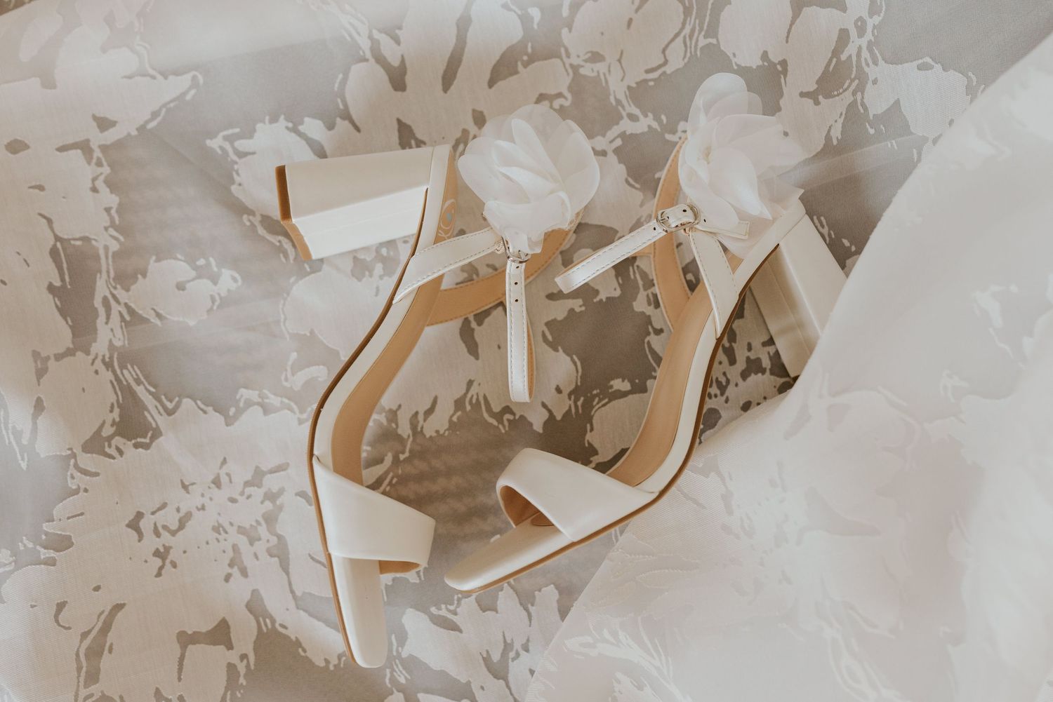 White bridal heeled sandals with ankle straps rest on a delicate lace wedding dress.